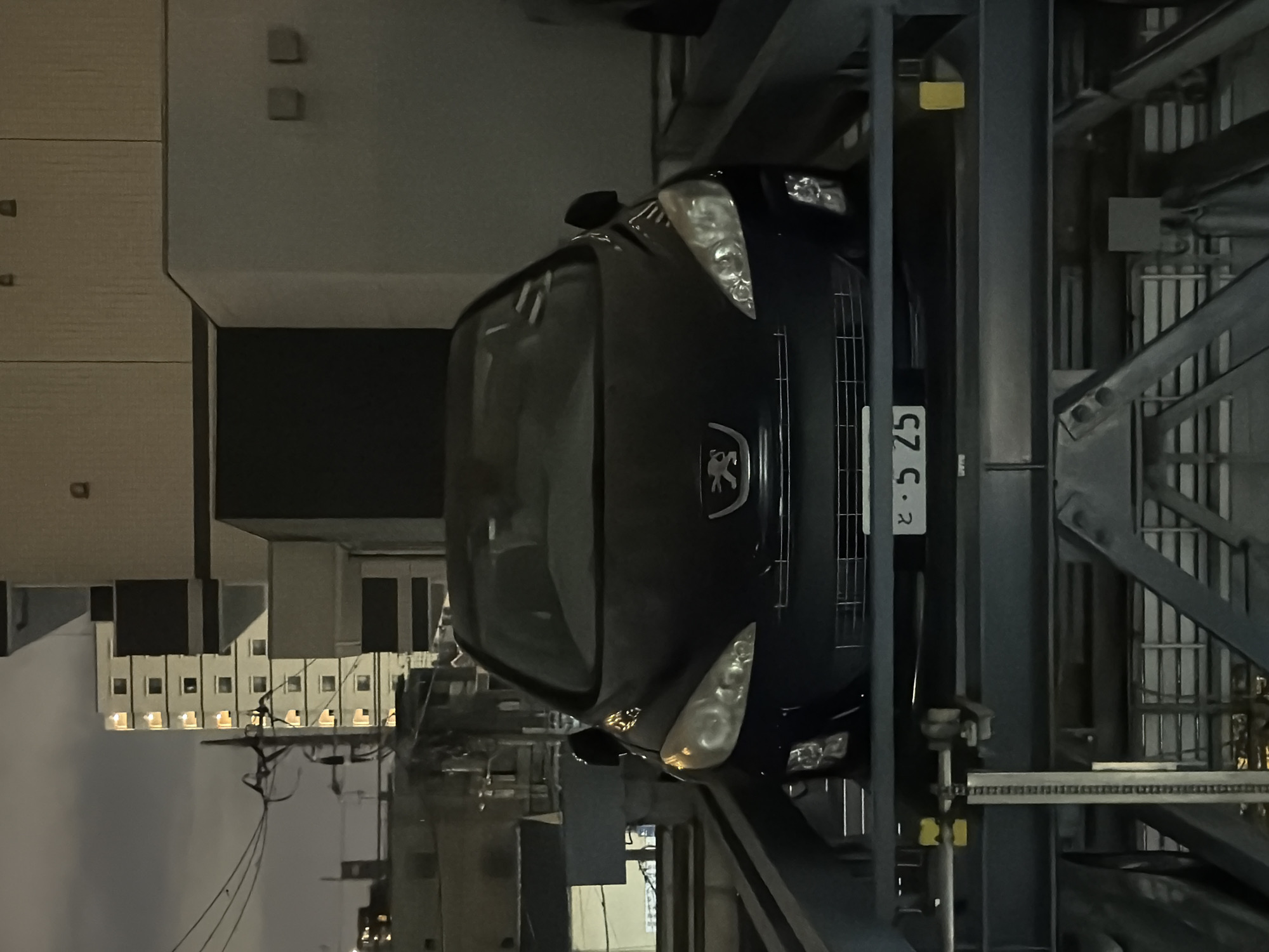 An elevated parking structure in Adachi, Japan, contains a dark-colored Peugeot car on a mechanical lift. The car faces forward, and its headlights are on. A license plate with the characters "と・5 25" is visible on the front of the vehicle. The car is positioned within the metallic structure of the parking lift. In the background, a multi-story building with illuminated windows is visible against a cloudy sky. The overall scene is dimly lit, suggesting it is nighttime.