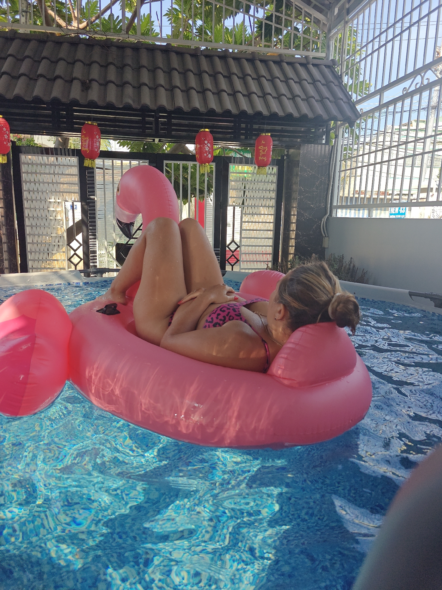 A woman is relaxing on a pink flamingo inflatable in a swimming pool. The pool is located outdoors, under a covered structure with a tiled roof. Red lanterns hang from the roof. The woman is wearing a leopard print bikini. In the background, a white fence and some buildings are visible. A sign with Vietnamese writing is attached to the fence. The water in the pool is clear and blue, with ripples on the surface. The overall scene suggests a leisurely day by the pool.