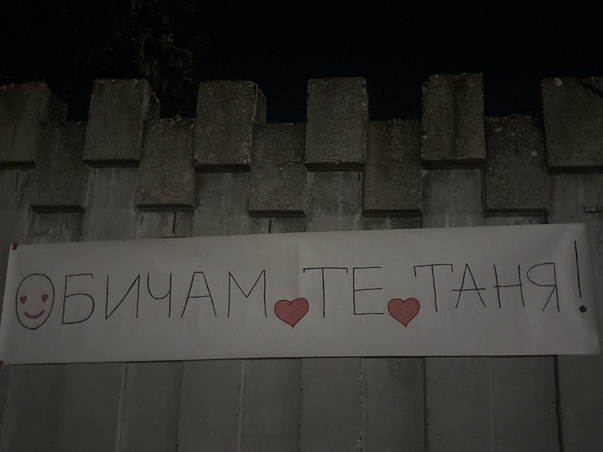 A handwritten sign in Cyrillic reads "ОБИЧАМ ТЕ, ТАНЯ!" which translates to "I love you, Tanya!". The sign features a smiley face with heart eyes and two pink hearts between "ТЕ" and "ТАНЯ". It is attached to a rough concrete wall or fence, with rectangular concrete blocks forming a textured background. The scene appears to be outdoors at night, with the dark sky visible above the wall. The concrete structure suggests an urban or semi-urban environment, possibly a park, public space, or a boundary wall in Veliko Tarnovo, Bulgaria. The lighting is low, casting shadows and highlighting the texture of the concrete.