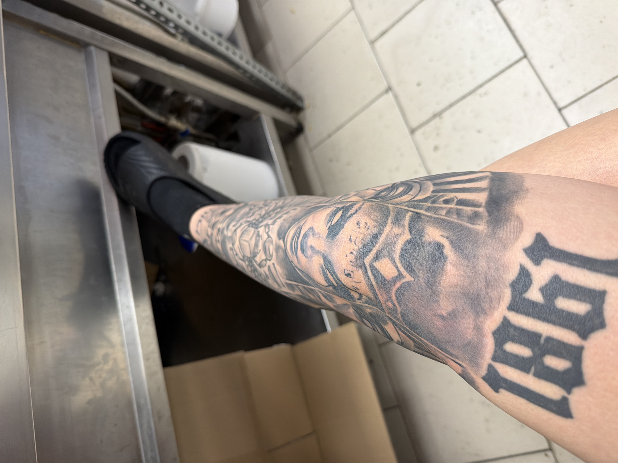 An indoor, eye-level shot captures a person's tattooed lower leg and foot, clad in a black slipper, within what appears to be a commercial kitchen or utility area in Plovdiv, Bulgaria. The left side of the frame is dominated by a reflective stainless steel counter or sink, with pipes and a metal shelving unit visible behind it. A white paper towel roll is partially seen underneath the counter. The floor on the right features light-colored tiles with grey grout. A brown cardboard box is present on the tiled floor next to the person's leg. The leg is extensively covered with a black and grey tattoo, depicting intricate designs including what appears to be a stylized face or figure, architectural motifs, and bold text. The year "1981" is prominently visible on the lower part of the leg in a distinctive gothic font. The scene suggests a functional, possibly industrial, indoor environment.