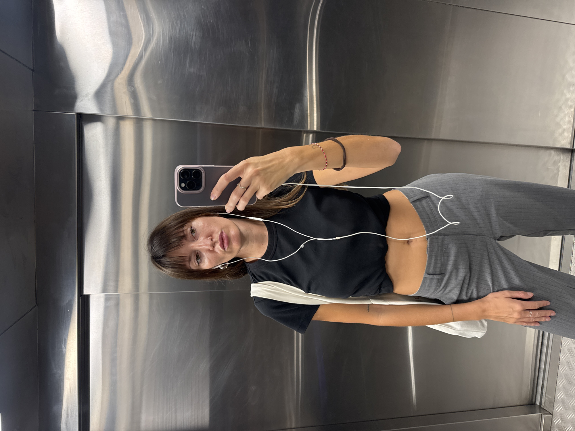 An indoor, eye-level, medium shot captures a woman standing in an elevator with brushed stainless steel walls. She is holding a smartphone in her right hand, taking a mirror selfie. She is wearing a black t-shirt, gray pinstripe trousers, and white earbuds. A white tote bag is slung over her left shoulder. She has brown hair with bangs and is wearing a ring on her left index finger and two bracelets on her right wrist. The elevator is well-lit, and the reflective surfaces of the walls create a slightly distorted view of the surroundings. The scene does not provide any specific clues about the city of Moscow, Russia.