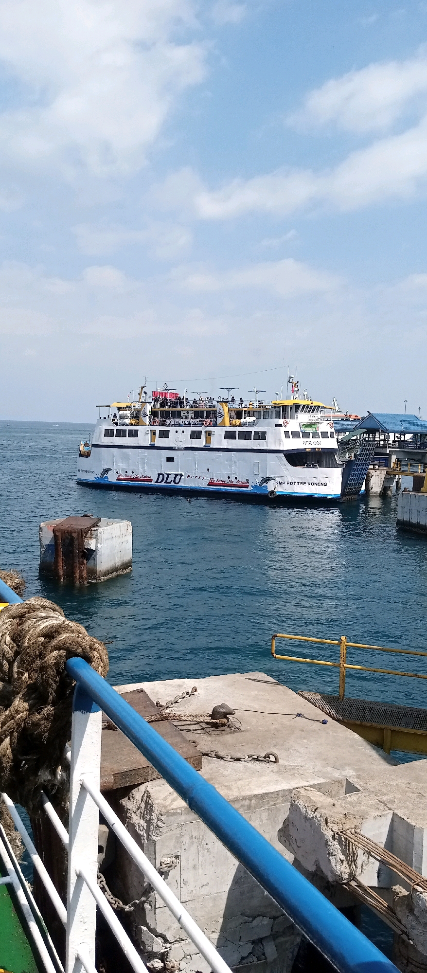 The image depicts a ferryboat docked at a pier in Banyuwangi, Indonesia. The ferry, named "KMP POTRET KONENG," is white with blue and yellow accents and has people visible on its upper deck. The pier is made of concrete, with thick ropes and chains securing the vessel. In the background, a blue-roofed structure and other docked vessels suggest a busy port. The water is calm, and the sky is partly cloudy. The scene is captured during daylight.