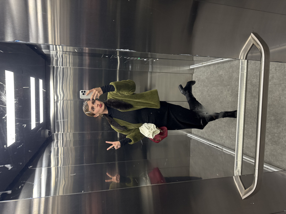 A person in an elevator takes a mirror selfie. They wear a green jacket with a fur collar, a black top and skirt, and black boots. A red and white handbag hangs from their shoulder. The person flashes a peace sign at the camera while holding a phone. The elevator walls are stainless steel, with a vertical grab bar visible in the reflection.