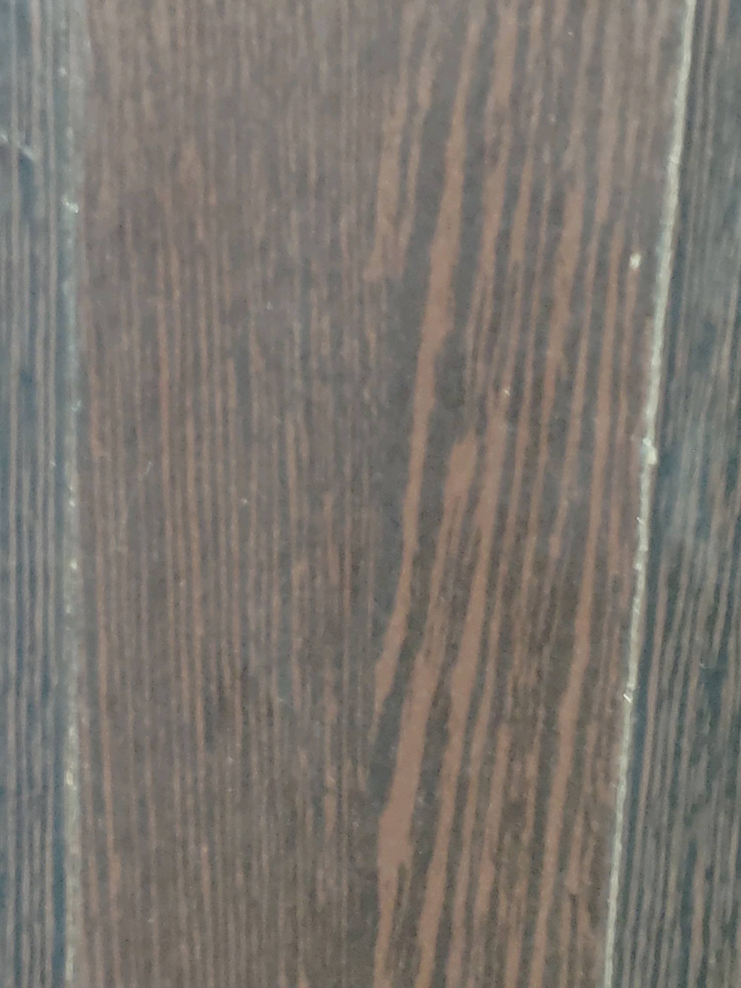 The image is a close-up, angled shot of a dark brown wooden surface, likely a floor or furniture. The wood grain is prominent, with vertical, wavy lines in varying shades of brown and black. There are subtle lighter streaks and occasional small, light-colored imperfections or dust specks visible on the surface. The lighting is somewhat dim, creating soft shadows that accentuate the texture of the wood. There are no people, animals, or other objects clearly identifiable in the frame. The image provides no information about its location in Jamshedpur, India, or any environmental context beyond the material itself.