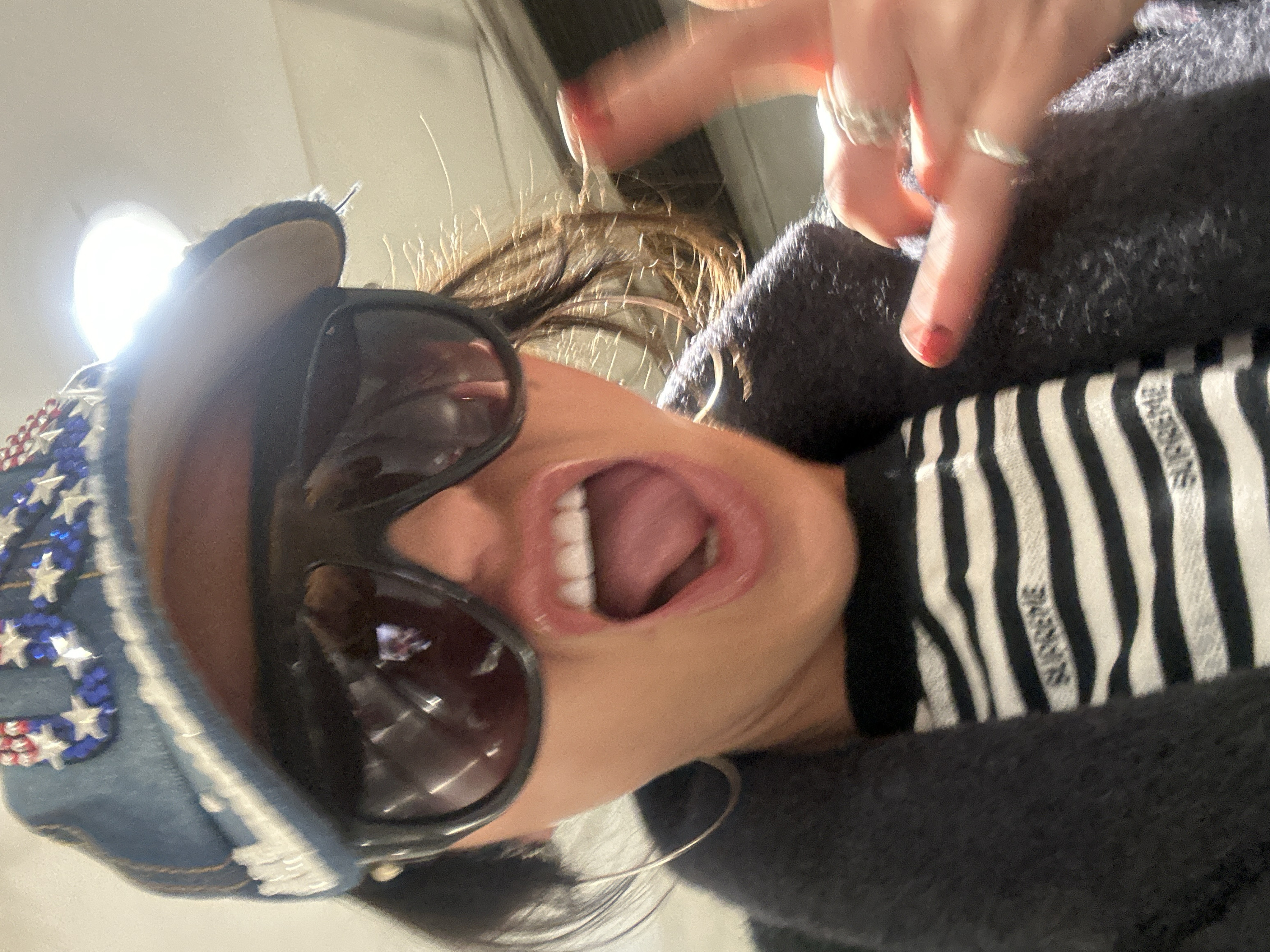 A person with dark sunglasses and a denim cap adorned with American flag-inspired embellishments. They are wearing a striped shirt with the word "SUPREME" visible. Their mouth is open, revealing their tongue and teeth. Their hand is visible in the upper right corner, with red nail polish and several rings. The image is angled, giving a distorted perspective of the subject.