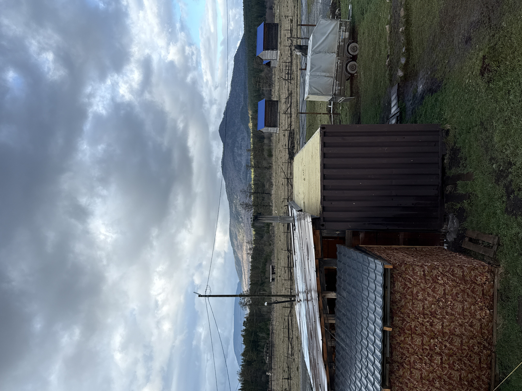 This image captures a rugged, rural landscape under an overcast sky, likely in Tyulyuk, Russia. In the foreground on the right, several structures are visible: a dark brown corrugated shipping container, an adjoining wooden structure with a light-colored metal roof, and a smaller shed with textured brown-patterned siding and a dark corrugated roof. To the left, a wooden utility pole with wires extends into the scene.
Beyond these foreground elements, a wide, dry, grassy field stretches towards distant mountains. Scattered across this field are several small, simple wooden shelters, each topped with a distinct bright blue roof. On the far right side of the field, a white-covered utility trailer with exposed wheels is parked.
The backdrop is dominated by a substantial mountain range, its lower slopes appearing forested. The sky is heavily clouded with varying shades of grey, indicating an overcast day, though some brighter patches suggest the sun might be partially breaking through. The ground in several areas appears damp or muddy, hinting at recent rain. No people, animals, or visible text are present, and no specific activity or event is taking place, presenting a still snapshot of a functional, utilitarian outdoor setting.