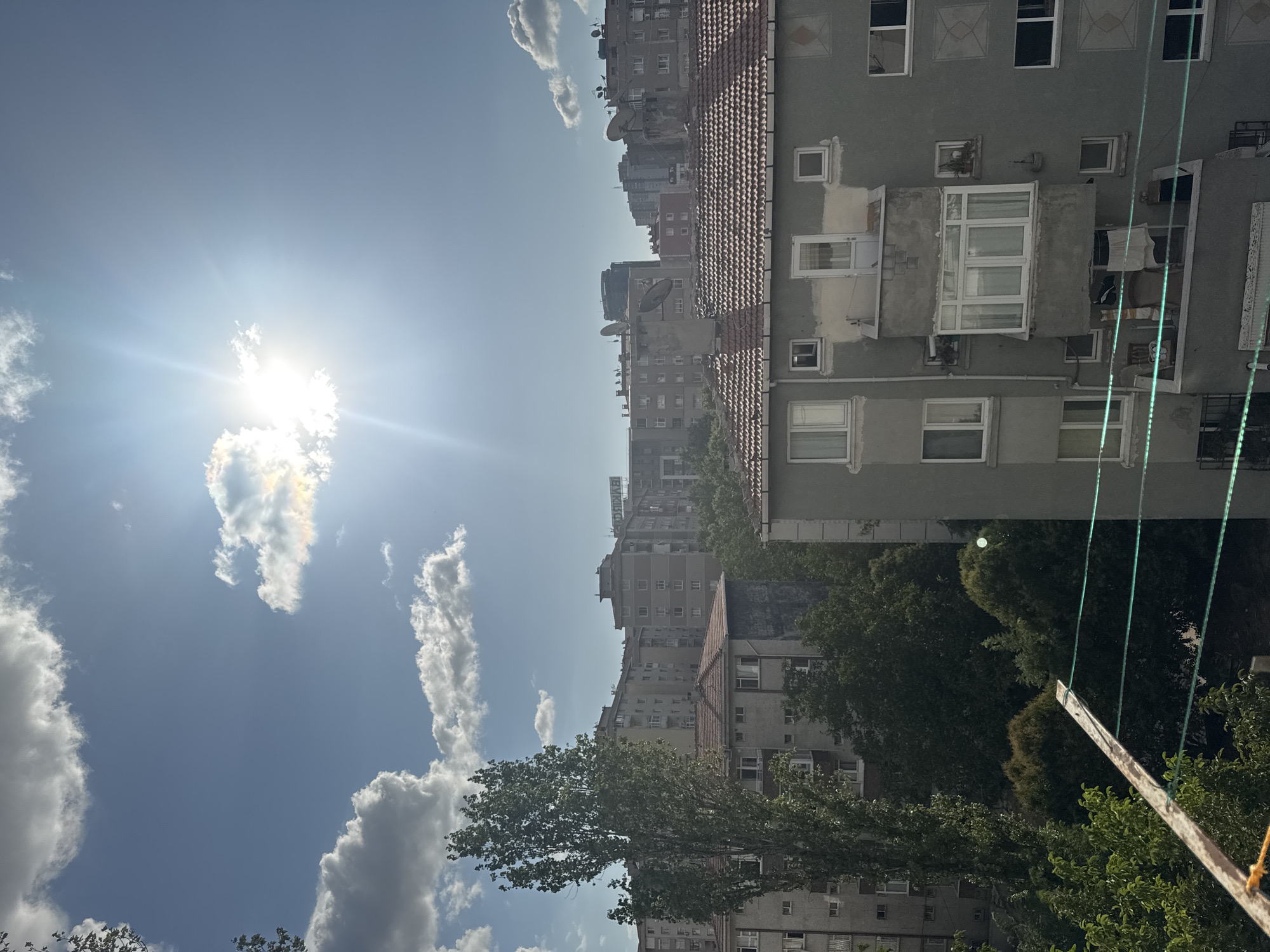 The image captures a view of an urban landscape in Gungoren, Turkey, under a bright, sunny sky with scattered clouds. The sun is prominently positioned high in the sky, casting lens flares and illuminating the scene. In the foreground, a part of a clothesline structure is visible, along with its green strings. The midground is dominated by a multi-story residential building with a tiled roof, featuring numerous windows and balconies. Behind this building, a dense collection of other apartment buildings, varying in height and architectural style, stretches across the horizon. Several trees, including a tall, leafy one on the left, punctuate the urban sprawl, their greenery contrasting with the concrete structures. Satellite dishes are visible on some of the rooftops, indicating residential areas. The overall impression is that of a typical day in a densely populated urban neighborhood in Turkey.
