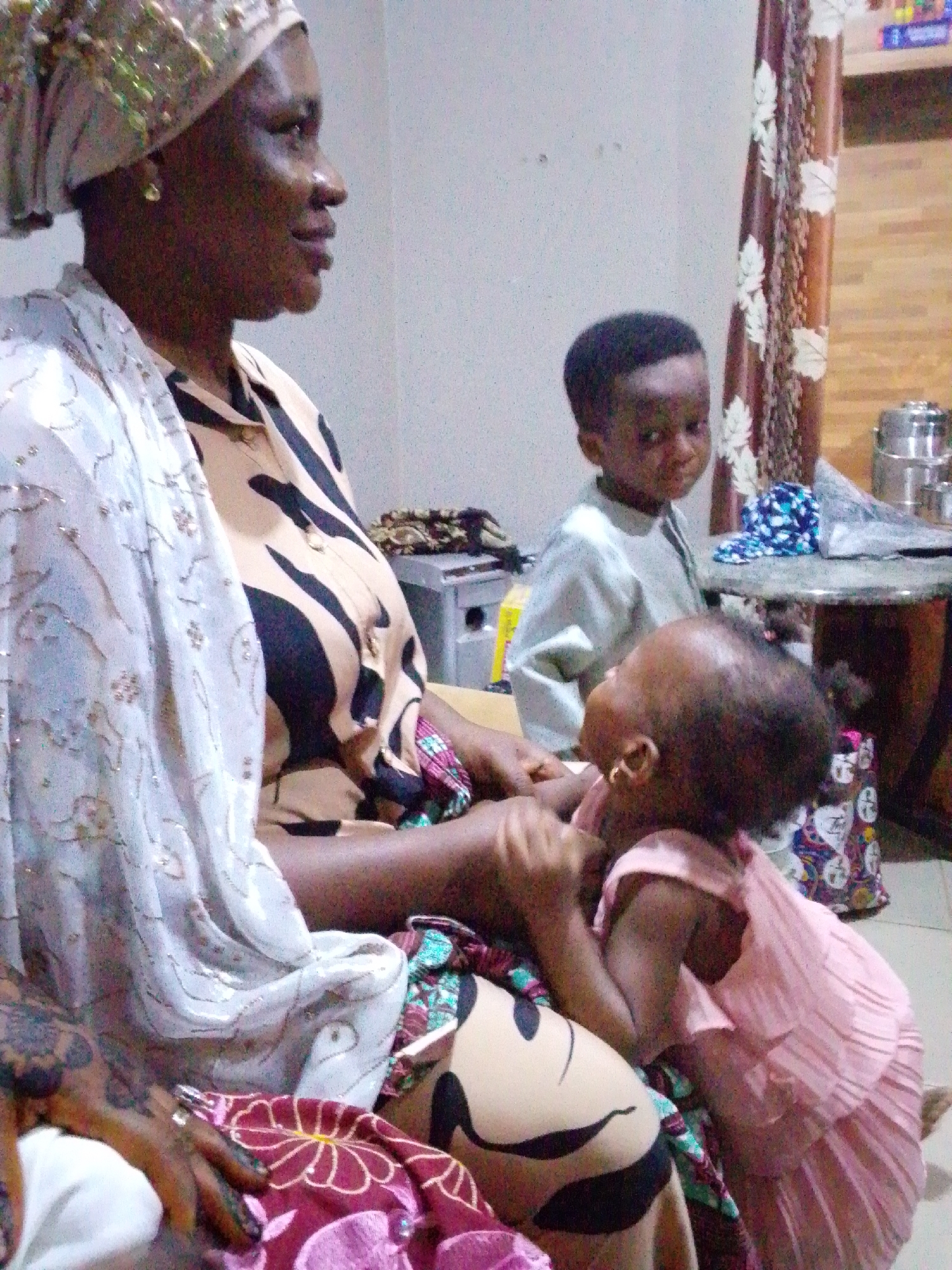 A woman is sitting, looking to the right, with a baby in a pink dress reaching up towards her face. A young boy stands behind the woman. The woman is wearing a patterned shirt and a head wrap, with a shawl draped over her shoulder. The setting appears to be indoors. Other objects visible include a curtain, a table, and some items on the table. The location is in the city of Unknown, Unknown.