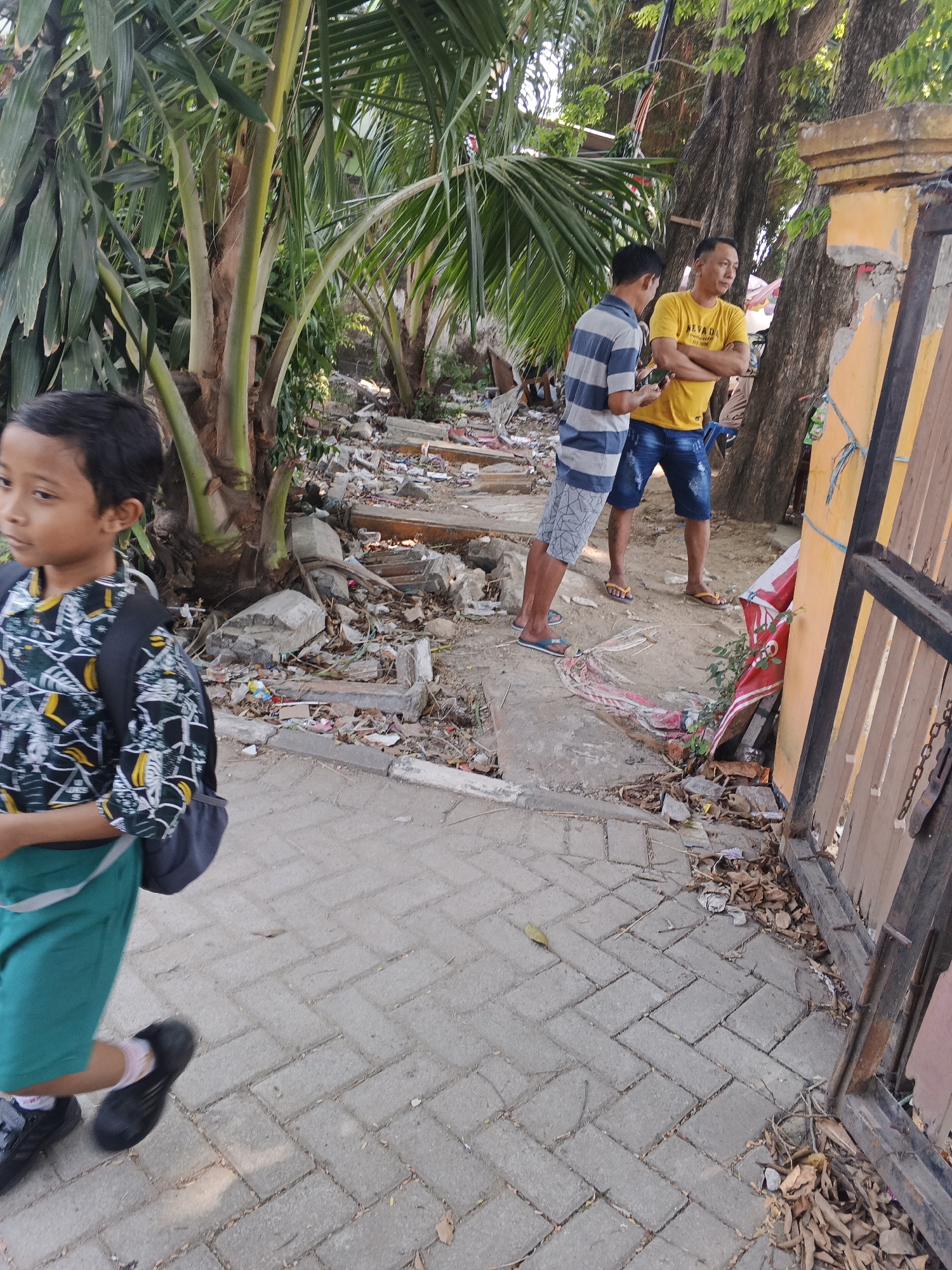The image depicts an outdoor scene, likely in Krian, Indonesia, featuring a young boy walking on a paved walkway. He is wearing a black and white patterned shirt, green shorts, a black backpack, and black shoes. Behind him, two men are standing near a large tree. One man, wearing a yellow t-shirt and blue denim shorts, has his arms crossed. The other man, in a blue and white striped shirt and patterned shorts, is holding a smartphone. The environment appears somewhat unkempt, with debris and construction materials scattered on the ground to the left of the walkway, adjacent to palm trees and other foliage. A weathered yellow wall and a dark metal gate are visible on the right side of the image. The lighting suggests it is daytime.