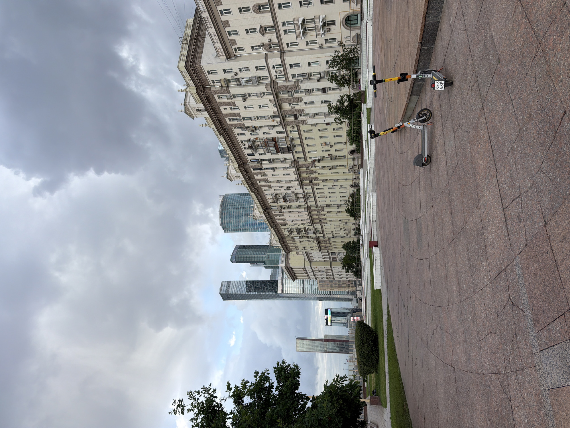 This image captures a wide outdoor view of Moscow City, Russia, under a cloudy sky. In the foreground, a textured red tiled plaza leads to a green grassy area with a neatly trimmed bush. Two electric scooters, one white with yellow accents and the other silver with yellow accents, are parked on the plaza. The scooter on the right has a license plate reading "KE3 63P".
Dominating the middle ground is a long, ornate, multi-story building with a light-colored facade, featuring numerous windows, balconies, and decorative architectural elements. Behind this building and to the left, a cluster of modern skyscrapers of varying heights and designs pierce the sky. Some of these skyscrapers have reflective glass facades, while one in the distance displays a bright digital advertisement. A large, leafy green tree is visible on the far left of the image, framing the scene. The overall atmosphere is one of urban modernity contrasted with classical architecture.