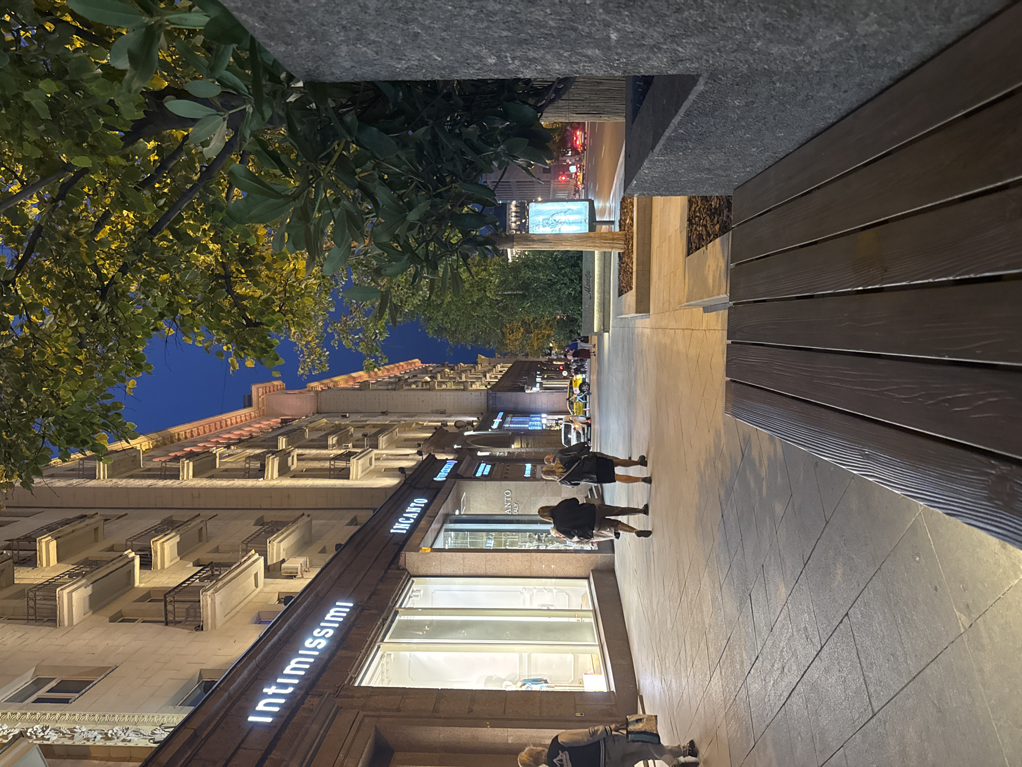 The image depicts a street scene at dusk in an urban environment. On the left, a multi-story building with neoclassical architectural elements lines the street, featuring balconies with wrought-iron railings. The ground floor is occupied by storefronts, including "Intimissimi" and "INCANTO." Pedestrians are visible on the sidewalk, including a woman in the foreground with blonde hair carrying a shopping bag. Further down the sidewalk, two women in dark clothing are walking away from the camera, and other figures are present in the distance. The sidewalk is paved with square tiles.
To the right of the sidewalk, there is a modern seating area with a wooden bench and stone planters with trees. Beyond the seating area, a street with vehicle traffic is visible, with car headlights and taillights illuminated, indicating it is evening. Trees with yellowing leaves line the street, and a digital billboard displays an advertisement. The sky is a deep twilight blue. The overall atmosphere is that of a city street as evening transitions into night.
