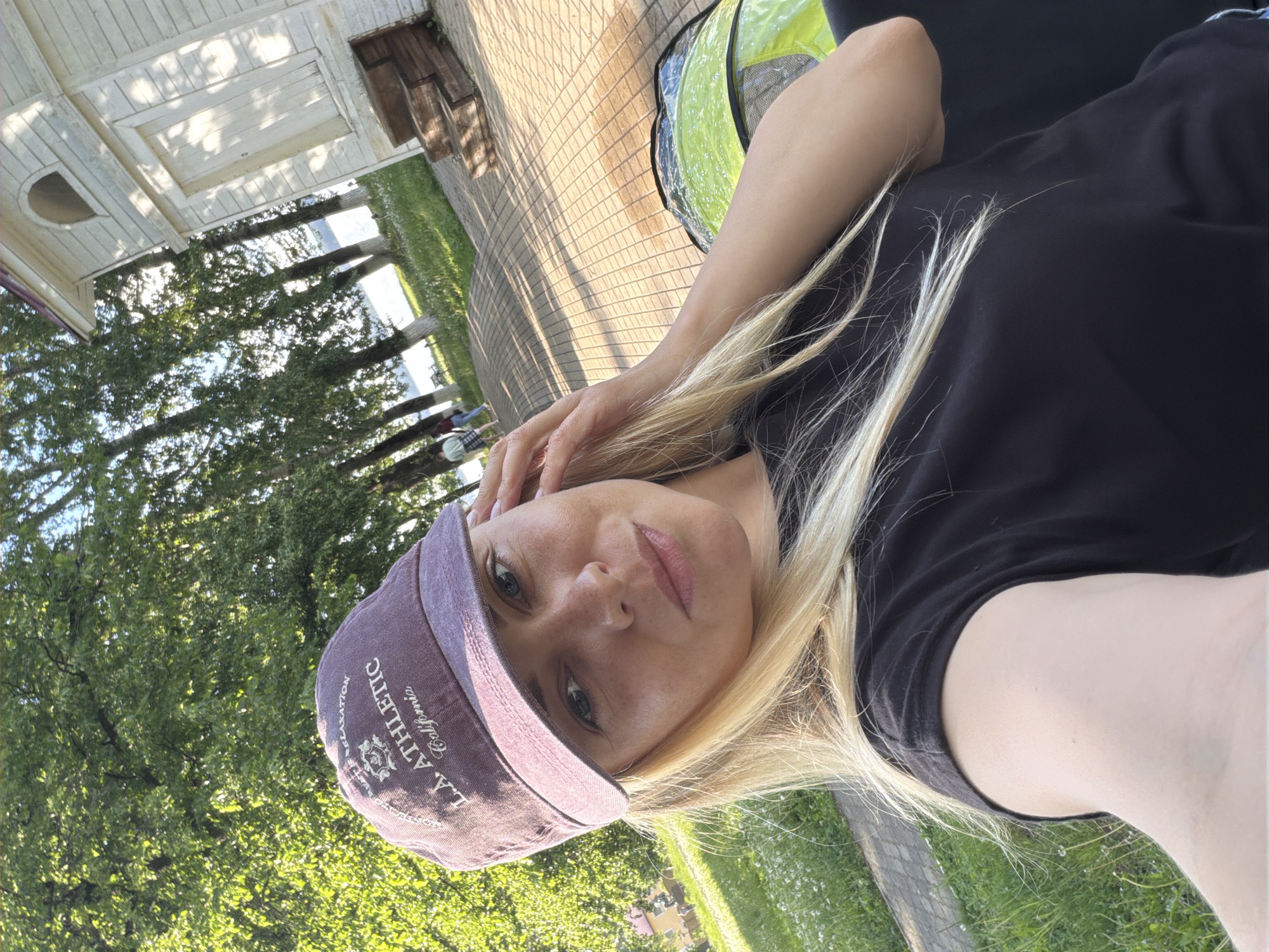 A young, fair-skinned woman wearing a purple baseball cap and a black T-shirt is taking a selfie outdoors in Votkinsk, Russia. Her blonde hair falls around her face and shoulder. In the background, trees and a body of water are visible. There is a white gazebo-like structure to the left. The ground is covered in grass and a brick pathway. The photo is taken at an angle, with the camera slightly above the woman. A yellow and green object lies near the brick pathway.