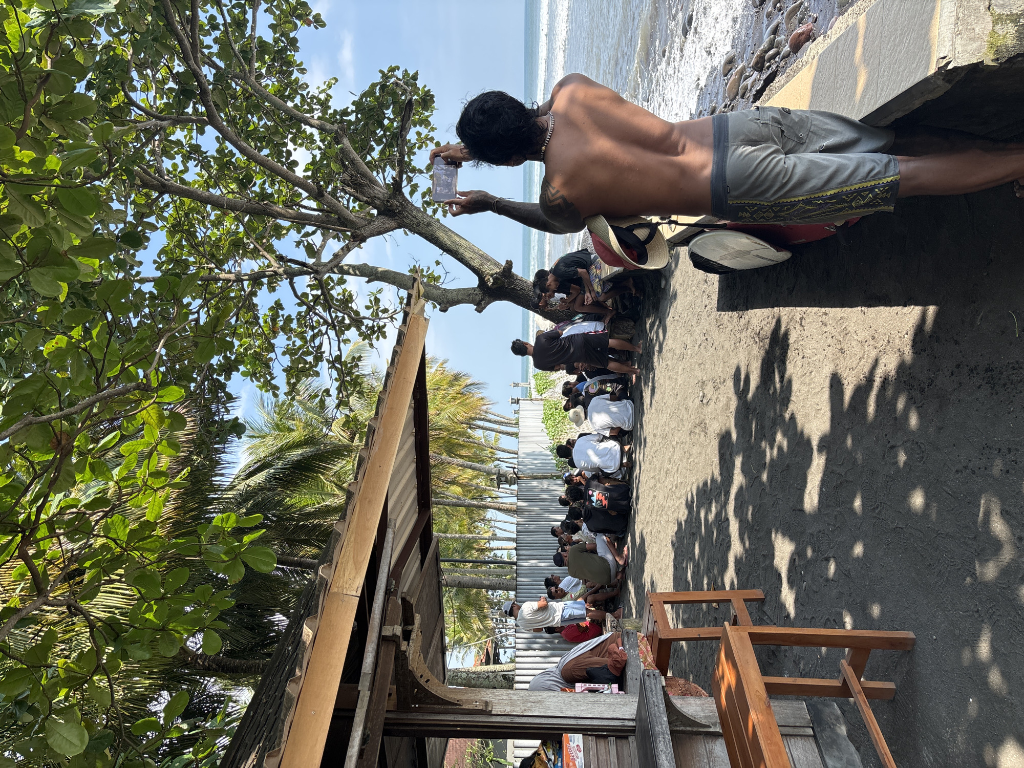 This vivid outdoor scene, likely set in Pekutatan, Indonesia, captures a group of people gathered at a dark sand beach under a bright, sunny sky. The image is oriented with the ocean and breaking waves on the right, and the land, featuring trees and structures, on the left.
In the foreground, slightly to the right, a bare-chested man with dark hair and a beaded necklace is seen from behind, leaning forward as he holds a smartphone to take a photo. He is wearing grey shorts with a patterned trim and has what appears to be a tattoo on his upper arm. Next to him, a red scooter with a white surfboard attached and a straw hat resting on it is visible.
Along a low concrete barrier separating the beach from an inner area, a line of approximately twenty people are seated on the dark sand. They appear to be looking out towards the ocean or an event taking place there. Some individuals are wearing white or dark t-shirts, and their postures suggest they are either resting or intently watching something.
To the far left of the image, a partially visible wooden shack or simple structure with a corrugated roof stands amidst lush green foliage. Large tropical trees with dense leaves provide shade, casting dappled shadows on the sandy ground, especially noticeable towards the bottom right. In the distance, a line of tall palm trees extends along the coastline.
The bright sunlight indicates it is likely midday or early afternoon, with a clear blue sky. The ocean water appears calm with gentle waves lapping the shore, which is composed of dark sand and some scattered rocks. No clearly discernible text is visible in the image. The overall atmosphere suggests a relaxed gathering or a local event by the sea.