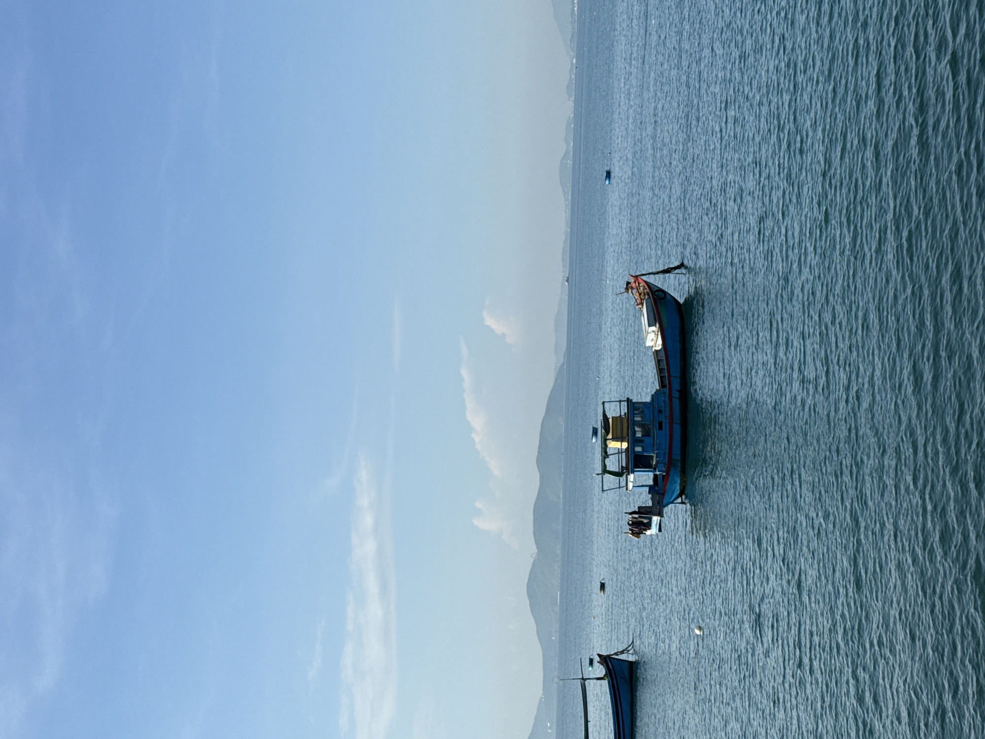 A scene of tranquil waters with several boats, likely in the waters off the coast of Nha Trang, Vietnam, under a clear blue sky with scattered white clouds. In the foreground, two blue and red fishing boats are anchored. One boat on the left is mostly visible, showing its bow and a mast. The boat on the right is more prominent, featuring a cabin structure with a yellow object on top and laundry hanging from its side. In the middle ground, a small white buoy floats on the surface. In the background, a chain of lush green hills stretches across the horizon, their peaks softened by a light haze. The ocean is a deep blue, with gentle ripples indicating a calm day. The overall impression is one of a peaceful maritime setting.