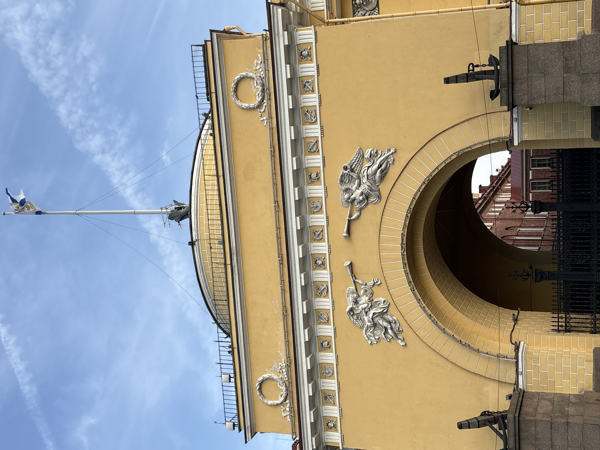 This is a low-angle, full-daylight outdoor shot of a yellow building with silver-colored architectural details. The building is identified as the General Staff Building in Saint Petersburg, Russia. The sky is blue, with light white clouds.
The building has a large archway through it, above which is a series of silver-colored sculptures of angels holding trumpets. There are decorative wreaths above the angel sculptures, and a row of square decorations lining the building.
A tall flagpole with a white and blue flag extends out from the roof of the building. A black metal anchor protrudes from the right side of the building, next to the archway. Through the archway, a red building can be seen. A black gate is in the lower right corner of the image.