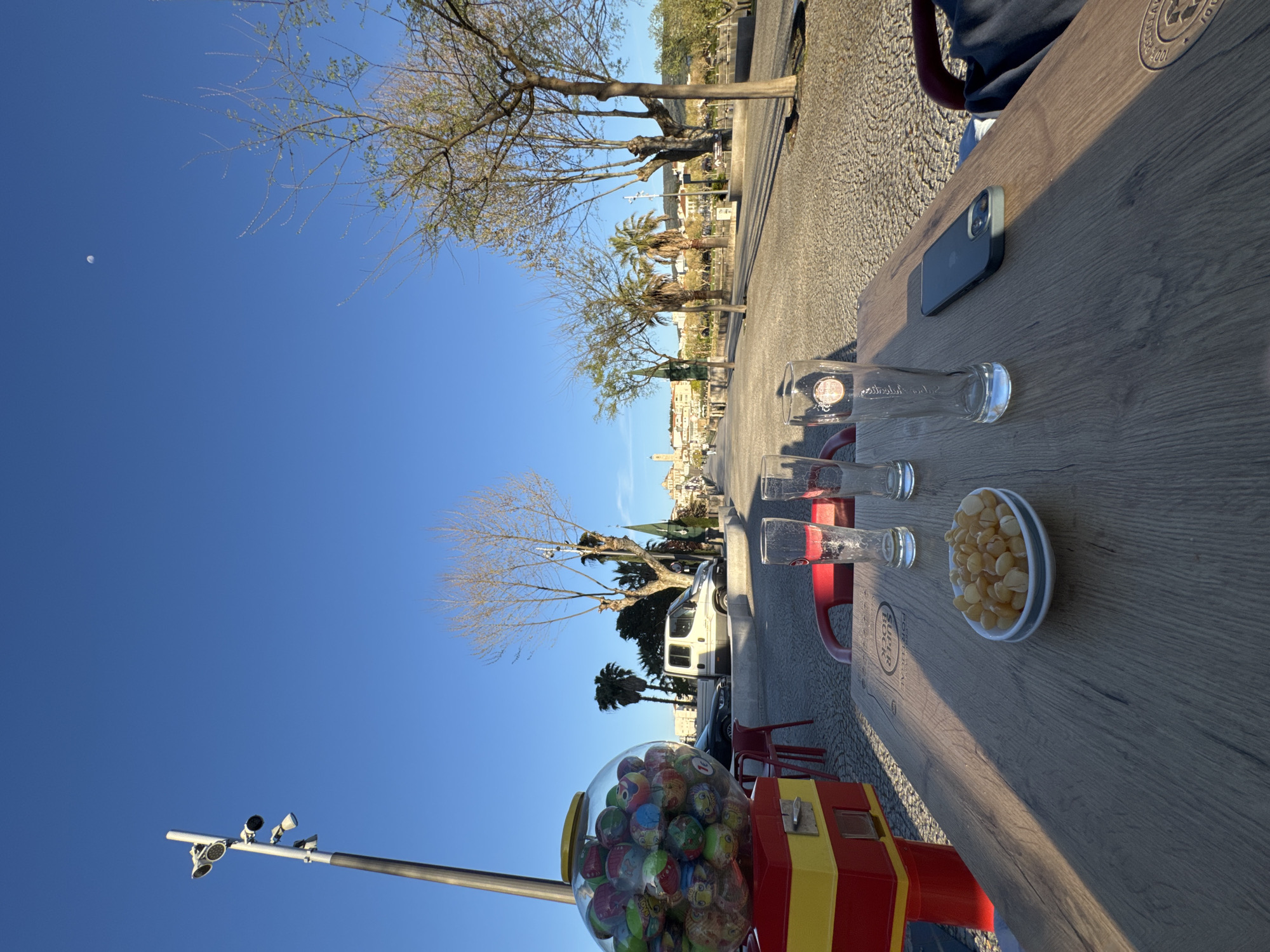 The image depicts an outdoor cafe setting along a paved promenade in Mirandela, Portugal, under a clear blue sky. In the immediate foreground, a light-colored wooden table is present, featuring a white oval bowl filled with light-colored snacks, three empty clear drinking glasses (at least one bearing a "Super Bock Portugal" logo), and a dark-colored smartphone lying face-down. Long shadows indicate a low sun angle. To the left of the table stands a red and yellow gumball machine filled with colorful capsules, adjacent to several red plastic chairs. A tall street light with multiple luminaires is positioned further left.
The midground reveals a wide, textured paved promenade extending into the distance, bordered by a mix of deciduous trees, some bare and some with nascent green leaves, alongside several palm trees. Parked vehicles, including a white van, are visible on a parallel street or parking area. The background features the distant cityscape of Mirandela, characterized by numerous buildings with varied roofs and at least one prominent tower or belfry. A distinct crescent moon is visible in the upper right portion of the clear blue sky. No individuals are present within the frame.