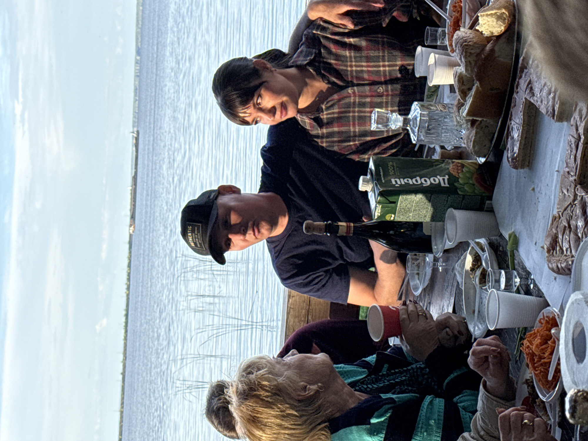 This is a medium shot, eye-level photograph taken outdoors. The scene depicts a group of people sitting around a table laden with food and drinks, with a body of water and a hazy sky in the background.
In the foreground, a table is covered with various dishes, including bread, sliced pastries, a salad, and what appears to be shredded carrots. There are also several paper cups and glasses, a bottle of wine, and a carton of juice.
Three individuals are prominently featured. On the left, an older woman with blonde hair is seated, holding a red cup. She is wearing a teal and black striped jacket. To her right, a man wearing a dark blue t-shirt and a black baseball cap with "AUTHENTIC" written on it is seated. He has his arm around the shoulders of a woman on his right. This woman has dark hair with bangs and is wearing a red and brown plaid shirt. Her left arm is resting on the man's arm.
The background shows a wide expanse of blue water with faint vertical lines of reeds or tall grass emerging from it. A long, flat boat or barge is visible on the horizon, as well as some distant landforms. The sky is a pale blue with scattered white clouds. The overall lighting suggests it is daytime, possibly late afternoon or early evening due to the warm tones and shadows.