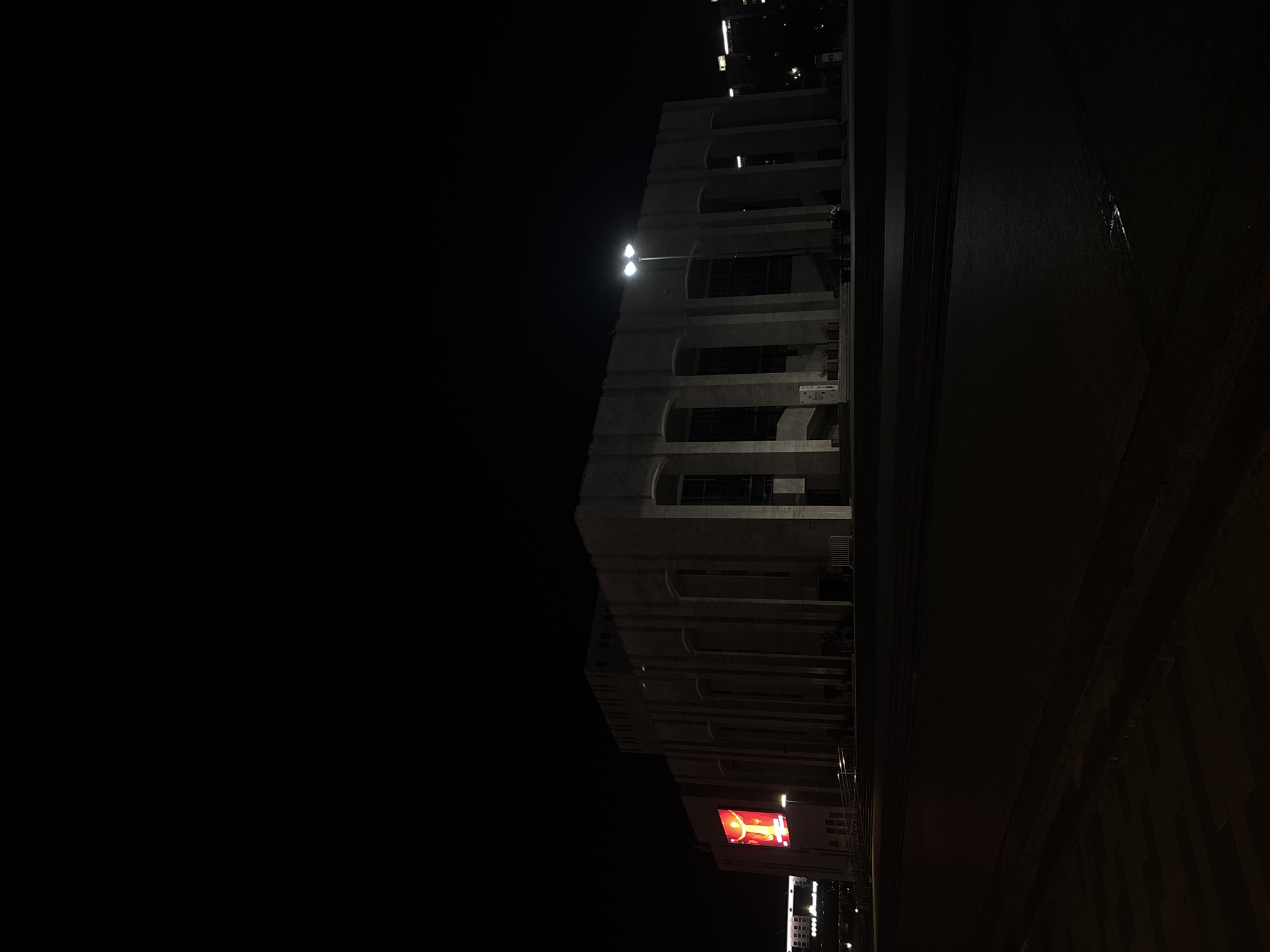 The image shows a nighttime view of a building in Perm, Russia. The building is a large, multi-story structure with a series of arched windows. A street lamp illuminates the building. A bright red sign is on the lower portion of the building. A street is next to the building and a street light is in front of the building. Other buildings can be seen in the background.