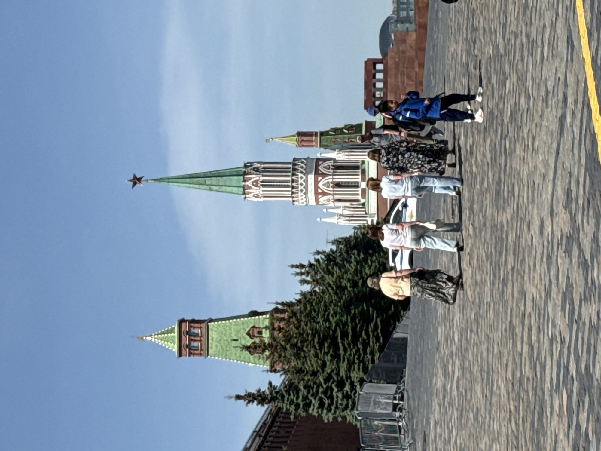 The image captures a segment of Red Square in Moscow, Russia, featuring the distinctive architecture of the Moscow Kremlin. Several individuals are visible, seemingly walking along the paved area. In the background, the Kremlin towers stand tall, identifiable by their unique design elements, including the star-topped spires. Lush greenery partially obscures the base of one tower. The sky is a clear blue. A yellow line is visible along the edge of the paved ground.