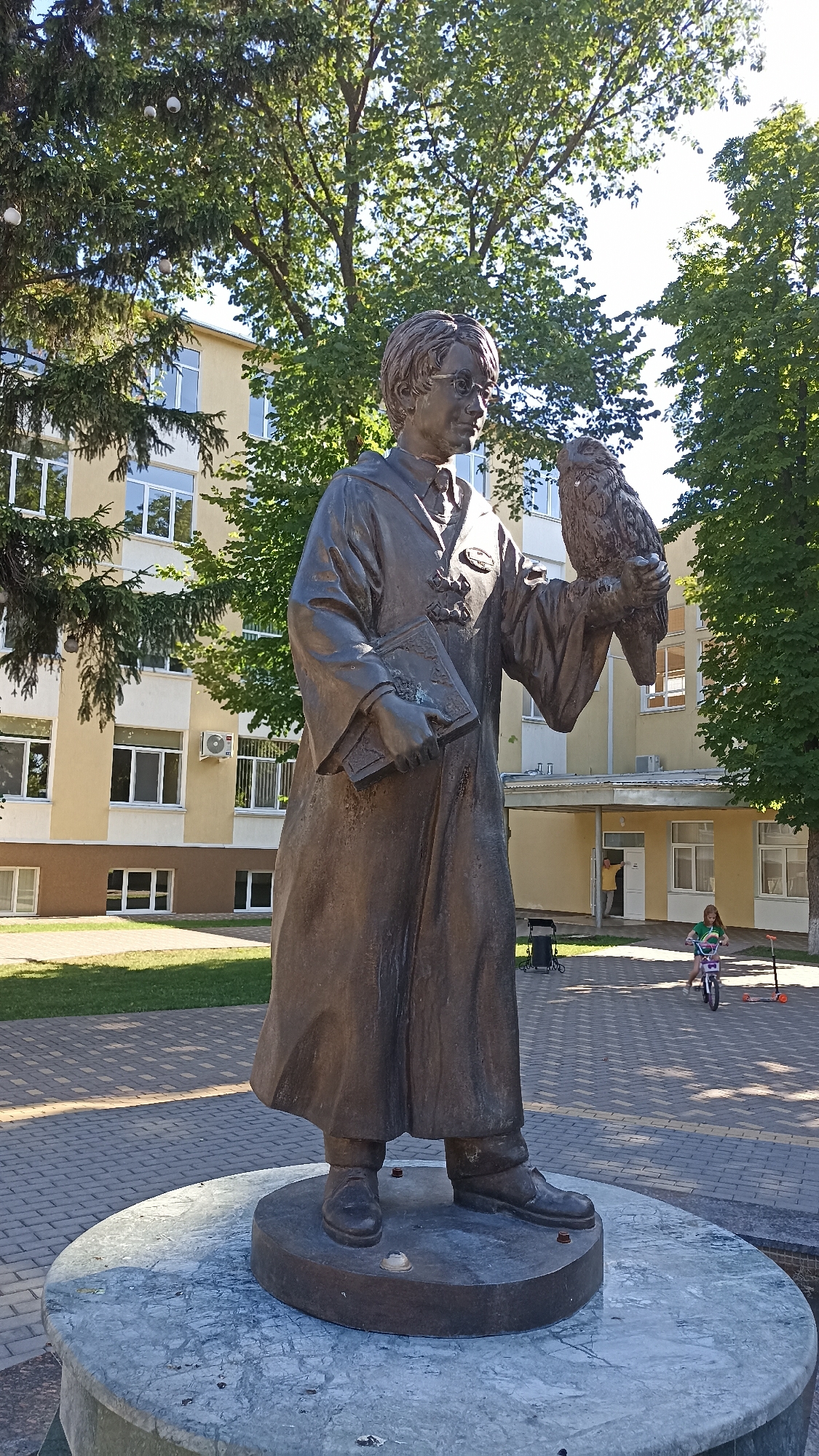 A bronze statue of a young boy with glasses and a robe is prominently displayed on a circular stone pedestal. The boy holds a book in his left hand and an owl in his right. The statue is located in Tiraspol, Moldova, in front of a pale yellow building with numerous windows. To the right of the statue, a young girl on a bicycle rides past, and a scooter is parked nearby. The scene is outdoors, with trees and dappled sunlight creating shadows on the paved ground.