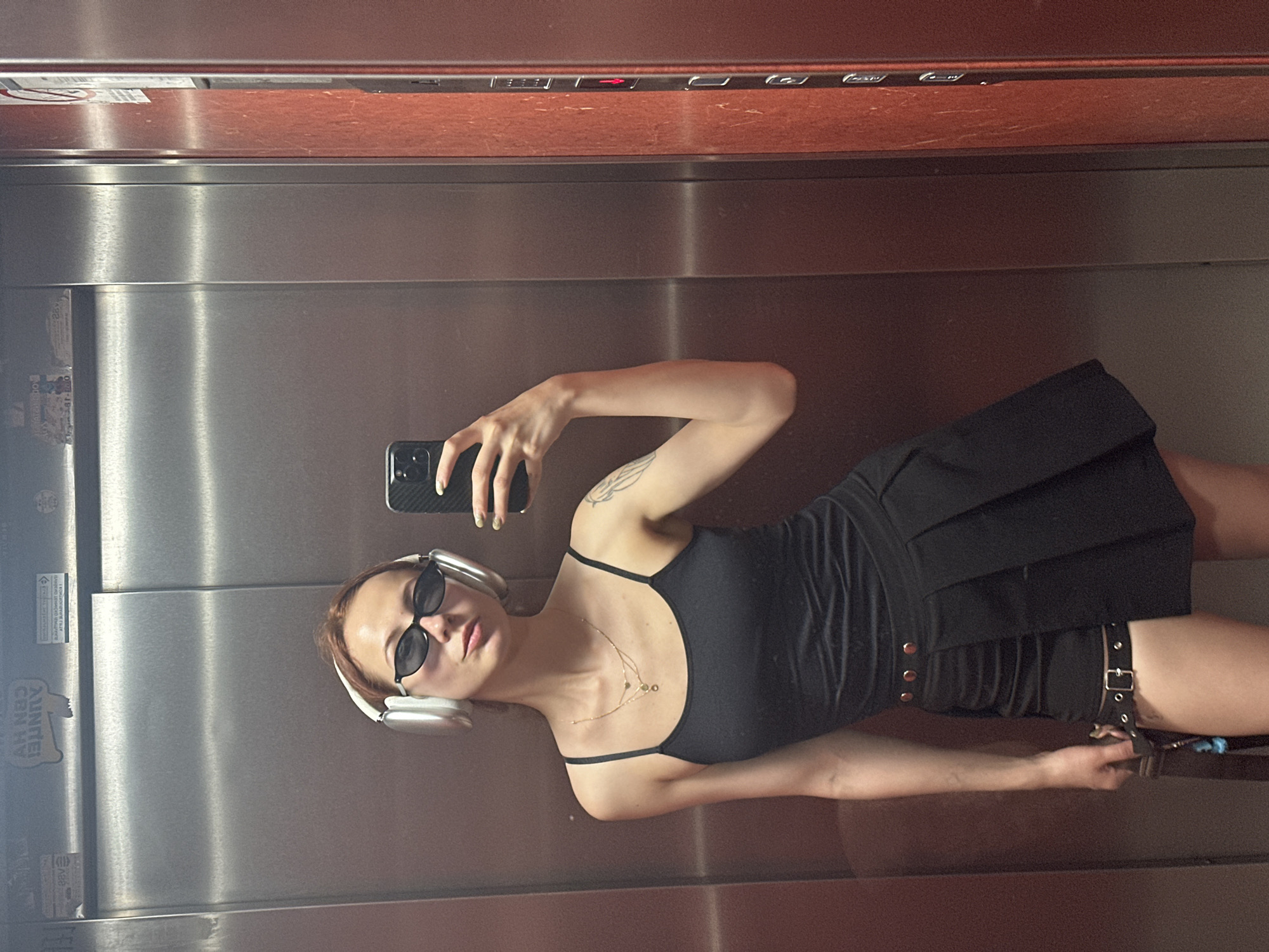 A woman takes a selfie in an elevator with metallic walls and a wood-paneled ceiling. She holds a smartphone in her left hand and wears sunglasses, white over-ear headphones, a black spaghetti strap top, a black skirt, and a layered gold necklace. The woman has a tattoo on her left arm. The location is in Novi Sad, Serbia. A small sticker is visible on the elevator wall.