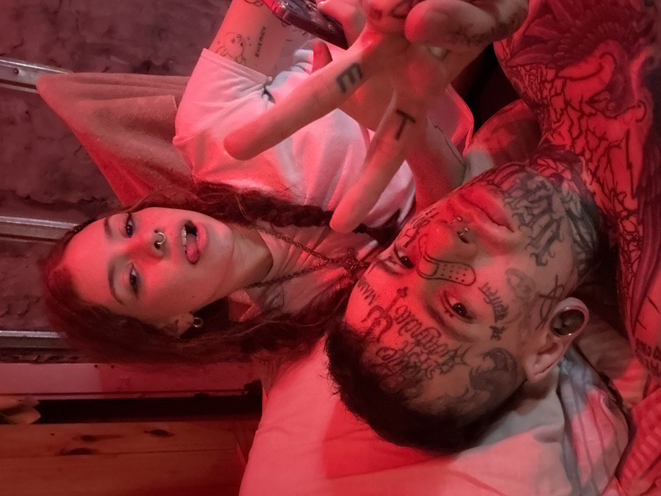 The scene depicts two individuals, a female and a male, in an indoor setting under dominant red lighting, likely a private residence within Buenos Aires, Argentina. The precise location (neighborhood or address) is not discernible from the visual information.
The female is positioned above and slightly behind the male, looking directly at the camera. She has long, dark hair styled in two braids, a septum piercing, and small hoop earrings. She wears a white short-sleeved t-shirt, revealing tattoos on her left arm, including what appears to be a stylized baby face and the word "ENERGY." Her left hand, adorned with additional finger tattoos (including "T" and "M"), holds a dark-colored smartphone and forms a "V" sign (peace sign). She is actively sticking out her tongue.
The male is lying down, positioned in the foreground, also looking towards the camera. He has extensive dark tattoos covering his entire face, neck, and upper chest, featuring various scripts, symbols, and a graphic resembling a band-aid on his right cheek. He has a nose piercing (septum) and large ear gauges/tunnels in both earlobes. His hair is dark and short.
Both individuals are in close proximity, appearing to be on a bed with white bedding visible in the foreground. The background is dimly lit and indistinct, showing dark, possibly textured wall surfaces. The red light casts a strong hue over the entire scene.