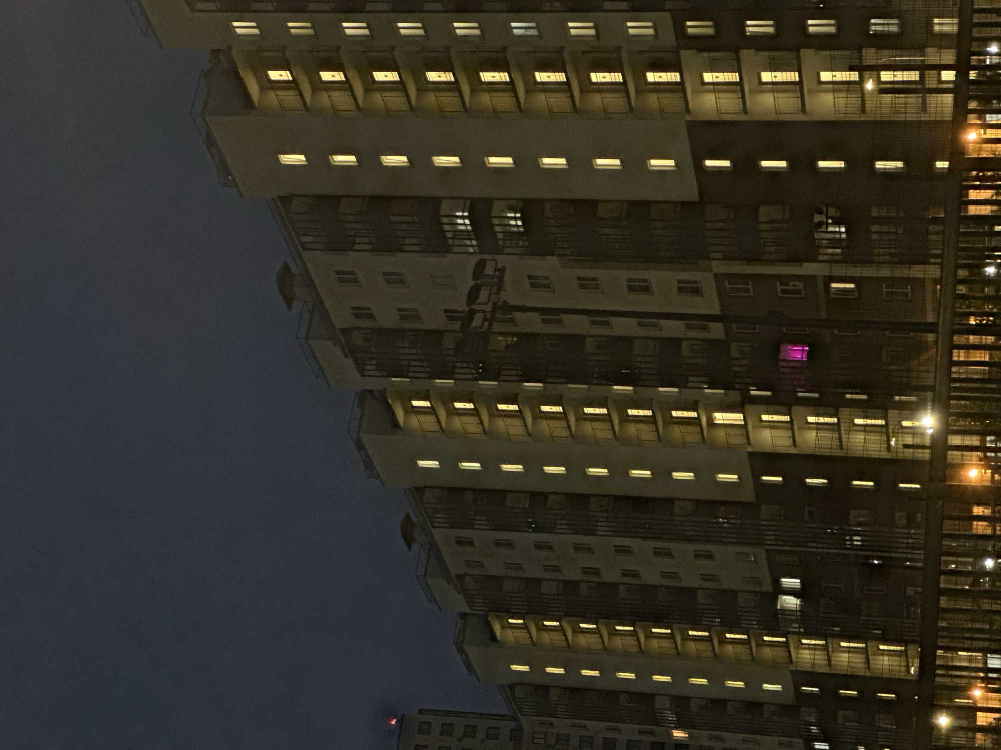 A low-angle, nighttime exterior shot captures a series of multi-story apartment buildings in the city of Unknown, Russia. The buildings, characterized by their beige and dark gray facades, are illuminated by numerous yellow lights emanating from their windows and balconies, suggesting occupancy. The sky above is a deep, dark blue, indicating it is late evening or night. A metal fence runs along the bottom of the frame, separating the viewer from the buildings and the street, where further lights are visible. The architecture features balconies with black metal railings on several floors. The overall scene depicts a residential urban environment after dark.
