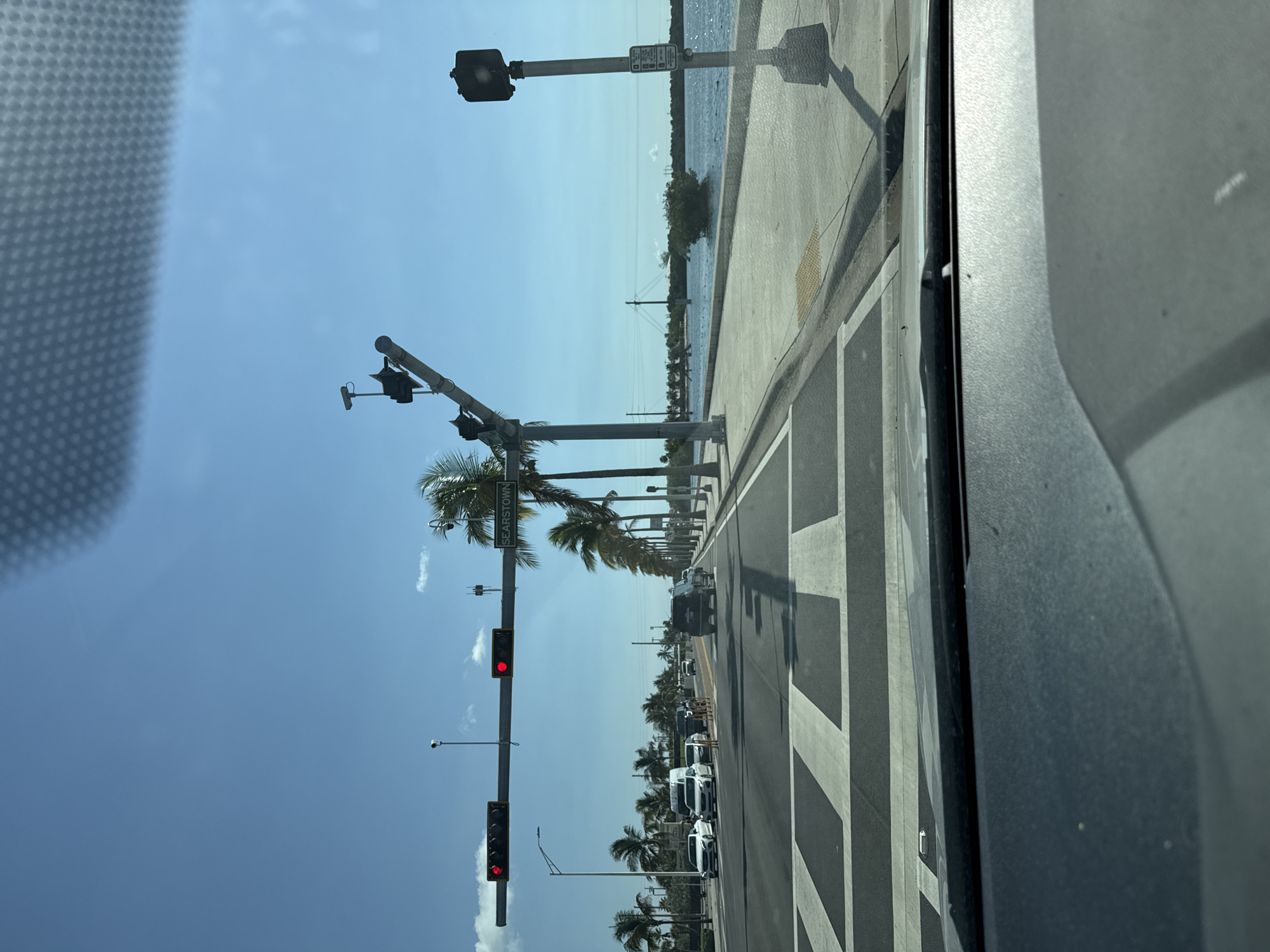 The image depicts a street scene in Key West, Florida, United States of America, on a clear, sunny day. The viewpoint is from inside a vehicle looking forward. A multi-lane road runs towards the horizon, bordered by a sidewalk and the ocean on the right. Traffic lights are visible, all displaying red, indicating a stop. Palm trees line the right side of the road, and a sign reading "SEARSTOWN" is attached to a traffic signal pole. Several vehicles are stopped at the intersection, including cars, a van, and an SUV. The road has a pedestrian crosswalk marked with white stripes. The ocean is calm and reflects the blue sky. In the distance, the water extends to the horizon, with some vegetation and structures barely visible. The foreground shows the dashboard of the vehicle from which the photo was taken.