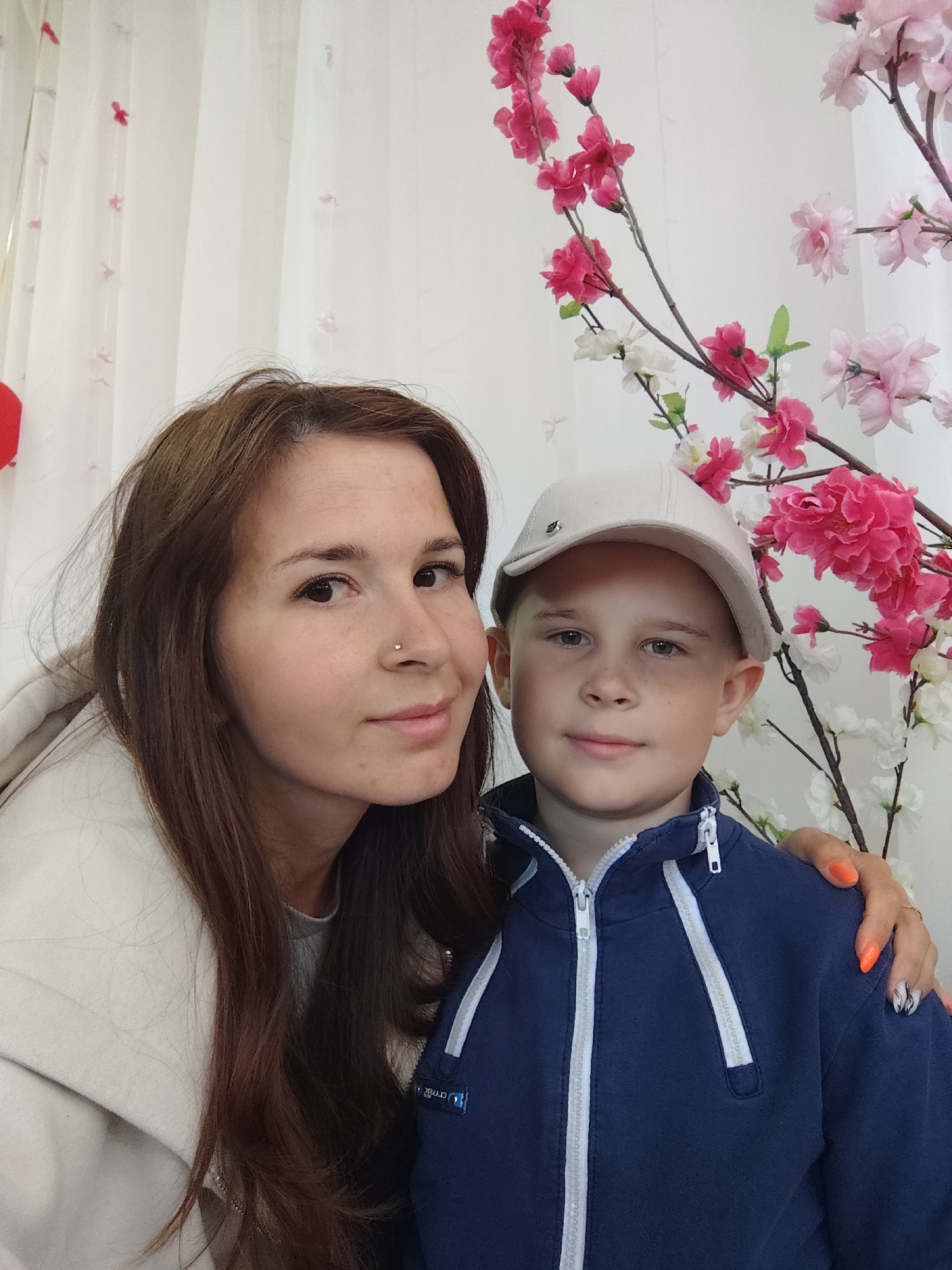 A medium shot depicts a woman and a boy posing for a photograph indoors in what appears to be a studio setting or decorated home environment. The woman, likely in her late 20s or early 30s, has light skin, brown hair, and a nose piercing. She wears a beige hoodie. The boy, likely around 8-10 years old, has light skin, short blond hair, and wears a light-colored baseball cap and a navy blue zip-up jacket with white trim.
The woman has her right hand on the boy's right shoulder. Both are looking toward the camera with slight smiles. Behind them is a backdrop consisting of white curtains or fabric, and decorative flowering branches with pink and white blossoms.
It's impossible to confirm the location in the city of Фоки, Russia, or any other place, because no other explicit geographical clues are visible.