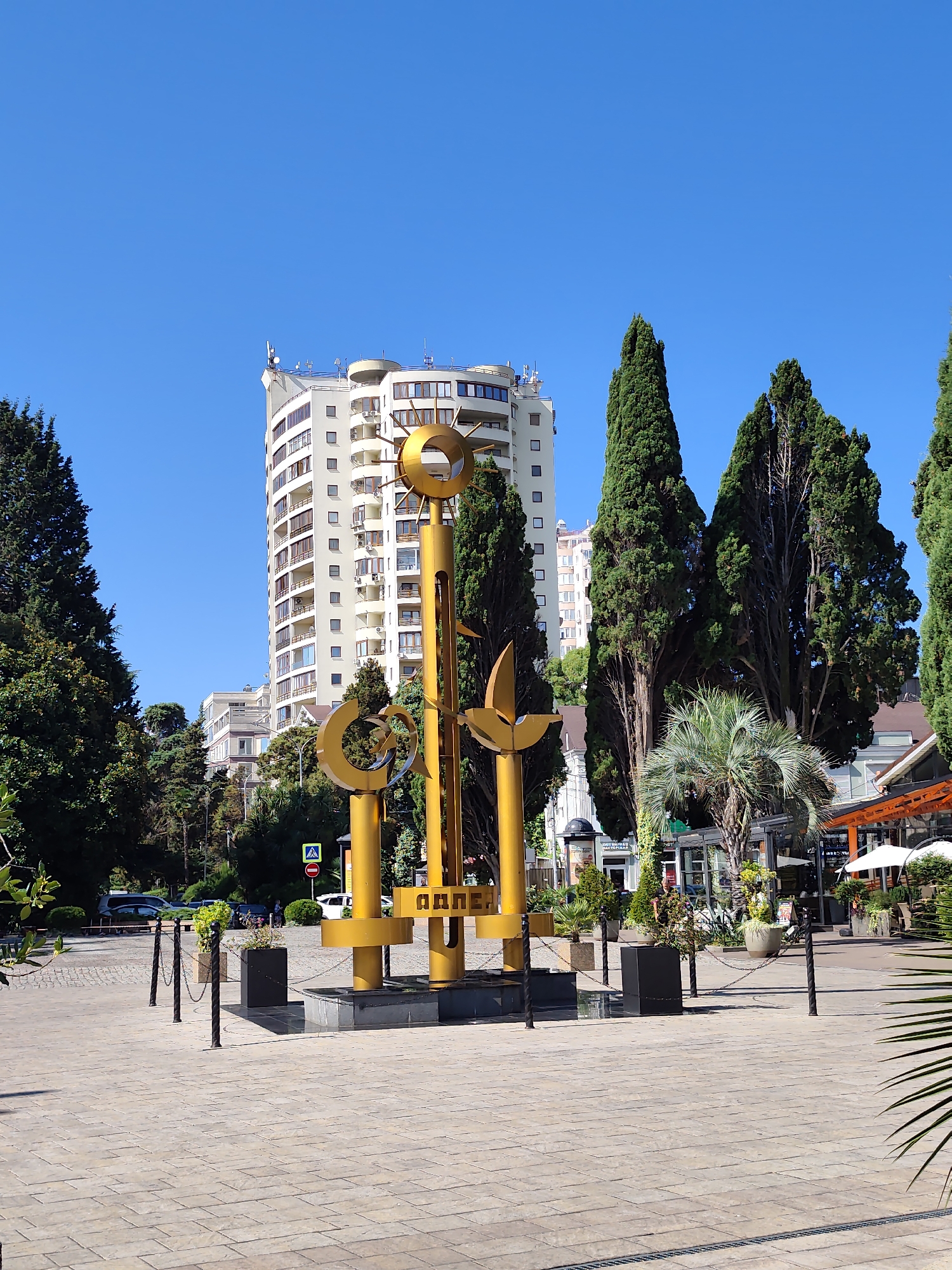This image depicts a sunny outdoor scene in Sochi, Russia, with a prominent golden sculpture in the foreground. The sculpture appears to be abstract, featuring geometric shapes and circular elements, possibly symbolizing the sun and other natural motifs. Behind the sculpture, a modern, multi-story building with a curved facade stands against a clear blue sky. Tall, slender cypress trees flank the building and the sculpture, adding a touch of natural greenery to the urban landscape. The ground is paved with stone tiles, and bollards with chains are placed around the sculpture, delineating a public space. In the background, there are glimpses of other buildings, possibly shops or cafes with outdoor seating, and parked vehicles. The overall impression is one of a well-maintained public area in a coastal city.