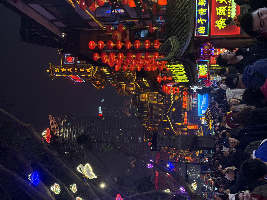 This image depicts a densely crowded street scene at night in Yuzhong District, China, likely within a commercial or tourist area such as Hongyadong. The environment is heavily illuminated by a combination of traditional and modern lighting elements.
**Buildings:** On the right, a series of multi-story buildings exhibit traditional Chinese architectural styles, characterized by dark tiled roofs with upturned eaves and multiple tiers. These structures are extensively adorned with yellow and white string lights outlining their architectural features. Numerous red Chinese lanterns are suspended in vertical rows along the facades, emitting a warm glow. Prominent neon signs, primarily displaying Chinese characters in red, yellow, and blue, are integrated into the architecture; for example, one sign high on a traditional building displays "鱼小不点" (Yú xiǎo bù diǎn), and another has the character "金" (Jīn) in gold. In the background, to the left of the traditional buildings, a modern, dark-colored high-rise skyscraper with numerous illuminated windows is visible, contrasting with the foreground's traditional aesthetic.
**People:** The foreground and midground are filled with a large crowd of individuals, primarily East Asian adults, walking and congregating. The density is high, with people occupying the entire visible street width. Individuals are dressed in various casual attire. No specific interactions or distinct actions beyond general pedestrian movement are discernible due to the crowd's density and the angle.
**Objects and Decorations:** Along the left side of the frame, large tree trunks are visible, decorated with illuminated butterfly-shaped lights in blue, red, yellow, and white. Red Chinese lanterns are a dominant decorative feature, appearing in multiple clusters. Various neon commercial signs are brightly lit, including a vertical sign on the far right that reads "桥头火锅" (Qiaotou Hotpot). A blue digital display screen is visible further down the street within the crowd. In the far distance on the left, faint vehicle lights suggest a street with traffic beyond the pedestrian area.
**Lighting and Atmosphere:** The scene is brightly lit by artificial sources against a dark night sky. The collective illumination creates a vibrant, bustling atmosphere. The overall impression is one of a lively urban setting, rich in cultural and commercial activity.