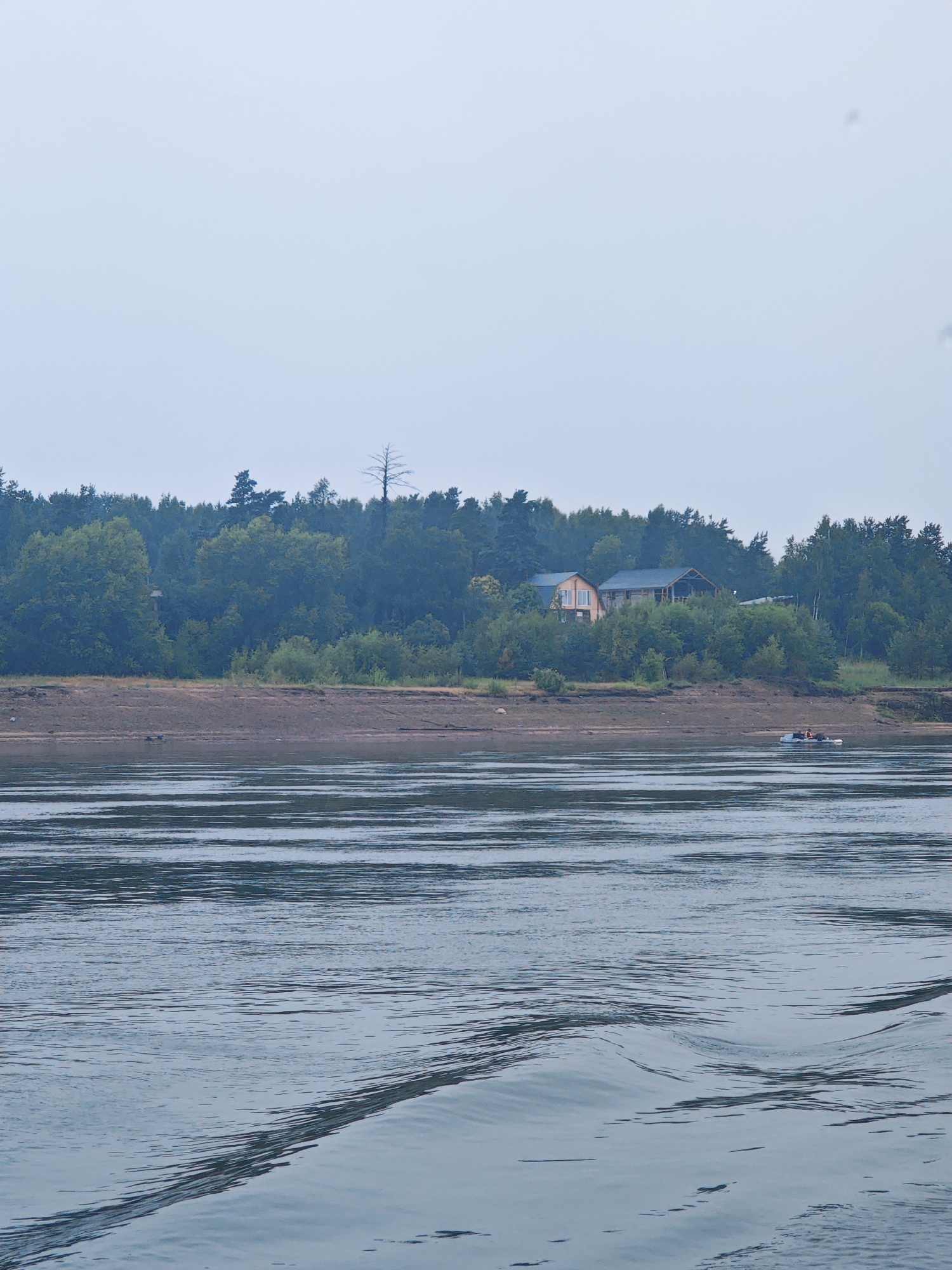 A wide, calm river flows in the foreground, with visible ripples and waves distorting the reflection of the overcast sky. On the far bank, a muddy shoreline is visible, topped by a dense forest. Nestled within the trees are two houses with wooden siding and dark roofs. In the middle distance, a small inflatable boat with two occupants is visible on the water near the shoreline. The scene appears to be in a natural setting, possibly a rural or semi-rural area on the outskirts of Lesosibirsk, Russia, given the presence of the river and forested landscape. The sky is a uniform pale gray, suggesting overcast or hazy conditions.