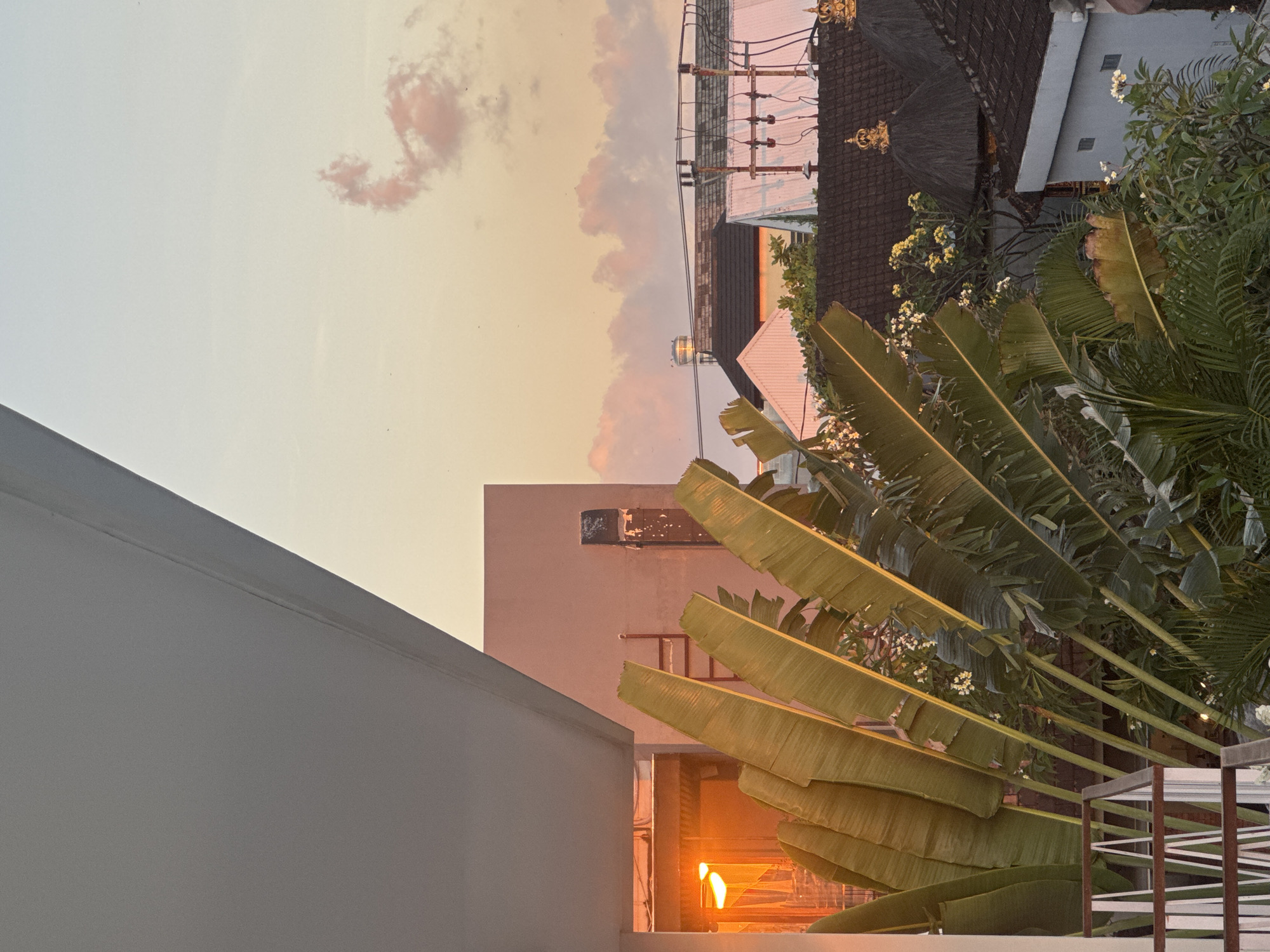 This is an outdoor, eye-level, medium shot captured at dusk in North Kuta, Indonesia. The foreground features large, green banana leaves that dominate the left and central portions of the frame. Behind the leaves, to the left, a building with an orange light emanating from an open window is visible. To the right of the banana leaves, rooftops of various buildings can be seen, some with dark, thatched roofs topped with golden crowns. Electrical wires and poles are also present amongst the rooftops. The sky is a soft gradient of peach and pale blue, with scattered, light pink clouds. No people are visible in the scene.