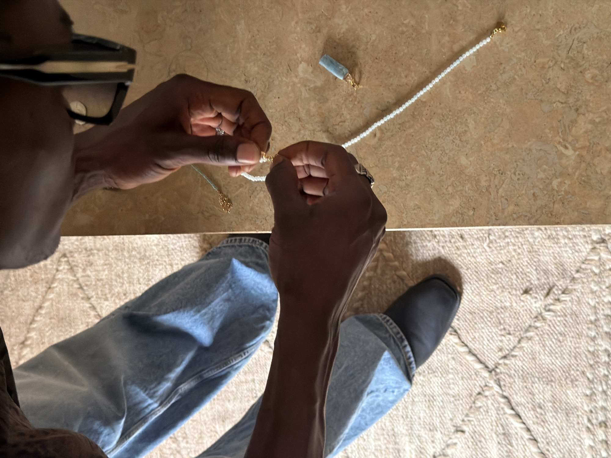 The image shows a close-up of a person's hands assembling a beaded necklace. The person is wearing blue denim jeans and black shoes, standing on a light-colored, textured rug. The hands are manipulating a white beaded string with a gold clasp and attaching a light blue rectangular pendant. The background consists of a beige, speckled tiled surface. The scene appears to be indoors, in a brightly lit area, possibly a workshop or a craft space. There is no indication of Marrakesh, Morocco in the image itself.