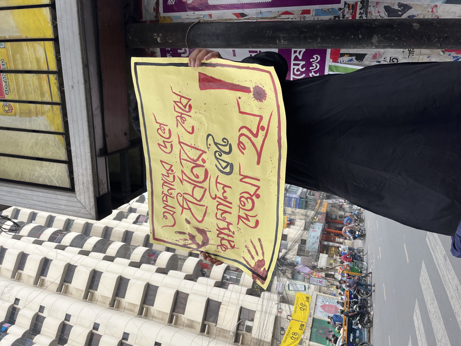 The image depicts an urban street scene in Dhaka, Bangladesh, dominated by a person holding a protest sign. The central figure is partially obscured by a rectangular yellow poster held at chest height. The person wears a dark, full-length garment, with only hands visible holding the sign. The sign features red Bengali text, reading "আপনাদের দেশে / ধর্ষণের শেষে / সম্মান কেড়ে / নেওয়া হয়" (translated as: "In your country / after rape / honor is taken away"). On the left side of the sign, there is a red handprint with splatter marks, and on the right, a large red exclamation mark also surrounded by red splatter.
In the foreground, an overhead metal structure, possibly part of an advertisement board or bus stop, extends from the right, partially visible above the sign. A section of this structure shows a partial advertisement in Bengali and English for "ADVANCED FORTIFIED SOYA OIL." A dark utility pole on the right is covered with numerous layered posters, predominantly purple and white, bearing Bengali script. The figure stands on or near a crosswalk, indicated by white stripes on the asphalt road.
The midground reveals a bustling street with a steady flow of traffic and pedestrians. Numerous motorcycles are parked or in motion, alongside several bicycle rickshaws carrying passengers or idling. Various individuals are visible on the street and sidewalks. The street is lined with multi-story buildings housing shops and commercial establishments at ground level, identifiable by colorful signage in Bengali. A dense network of overhead electrical wires crisscrosses the street.
In the background, a tall, light-colored building with multiple balconies and windows rises prominently on the left, indicative of a high-rise structure. The sky is bright, suggesting daytime conditions.