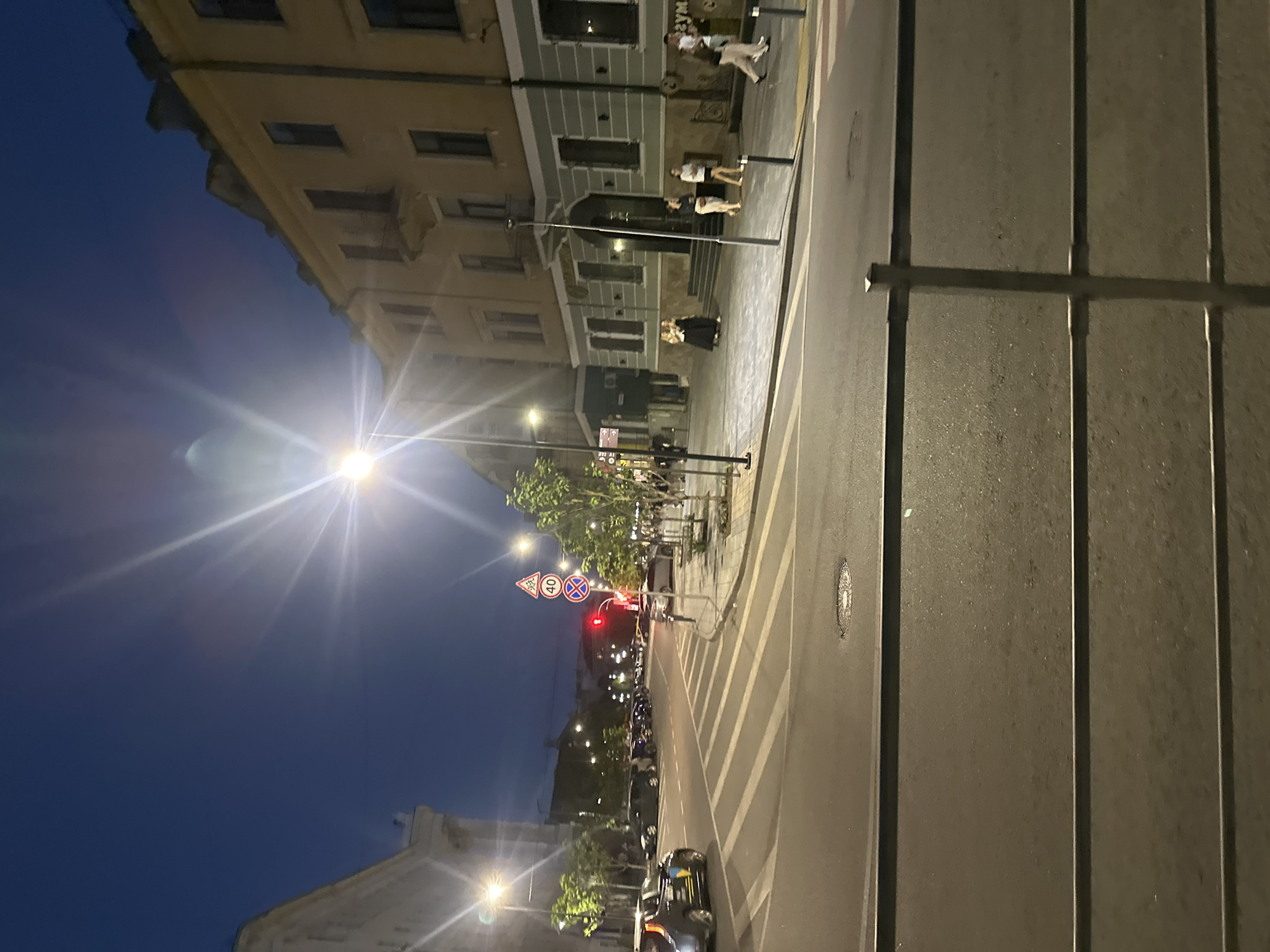 The image depicts a nighttime street scene in Ternopil, Ukraine. The perspective is from a street-level view, looking across a crosswalk towards buildings on the opposite side of the street. The sky is a deep blue, indicative of twilight or early night, with a bright streetlamp casting strong rays of light that cause lens flare.
On the left side of the street, a white building with ornate architectural details is visible. Cars are parked along the curb, and a dark-colored car, possibly a taxi with a Ukrainian flag decal, is in motion on the street.
Across the street, a yellow building with multiple stories dominates the right side of the frame. The building has numerous windows and a prominent balcony on the second floor. A group of people are walking on the sidewalk in front of this building. Two women in formal attire are standing near the entrance of the building, while other individuals are seen walking in both directions.
Traffic signs, including a speed limit sign showing "40" and a warning sign with a triangle, are visible near the intersection. A red traffic light is illuminated. Trees line the sidewalk, their foliage illuminated by streetlights. The street itself appears wet, reflecting the artificial light. A metal railing is in the foreground, partially obscuring the view of the street and sidewalk. A manhole cover is visible on the road surface. The overall impression is of a European city street at dusk or early night, with ongoing pedestrian and vehicular activity.