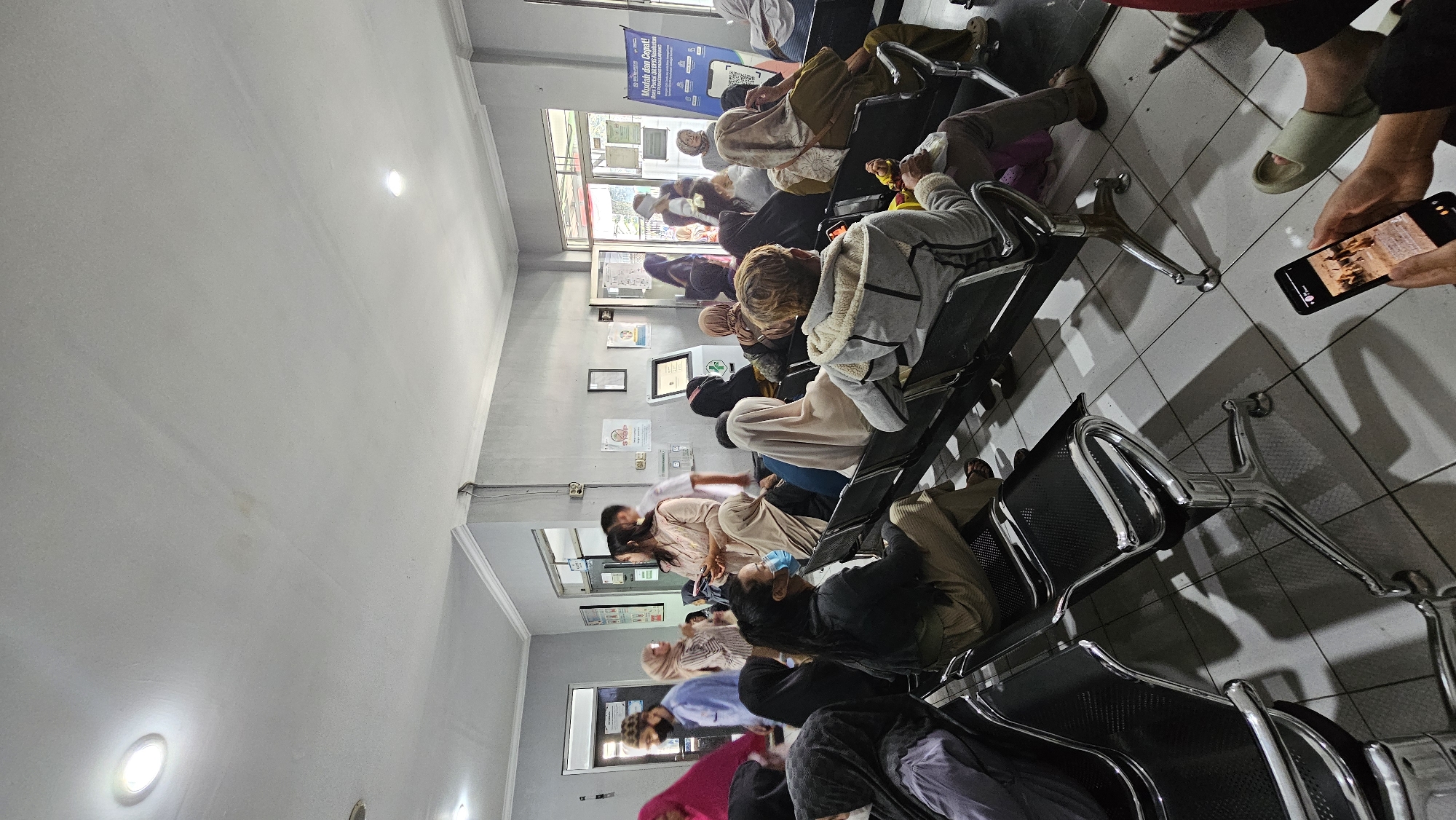 The image depicts an indoor waiting area, likely a clinic or hospital, with a tiled floor and grey walls. Several individuals are seated on black metal chairs, appearing to wait. The majority of people are wearing headscarves, and some are wearing face masks. The lighting appears to be overhead fluorescent lights. In the background, there are doorways and a digital kiosk or screen. A banner with text and graphics is visible near an open doorway. One person in the foreground is holding a smartphone displaying an image. The overall atmosphere suggests a public waiting space.