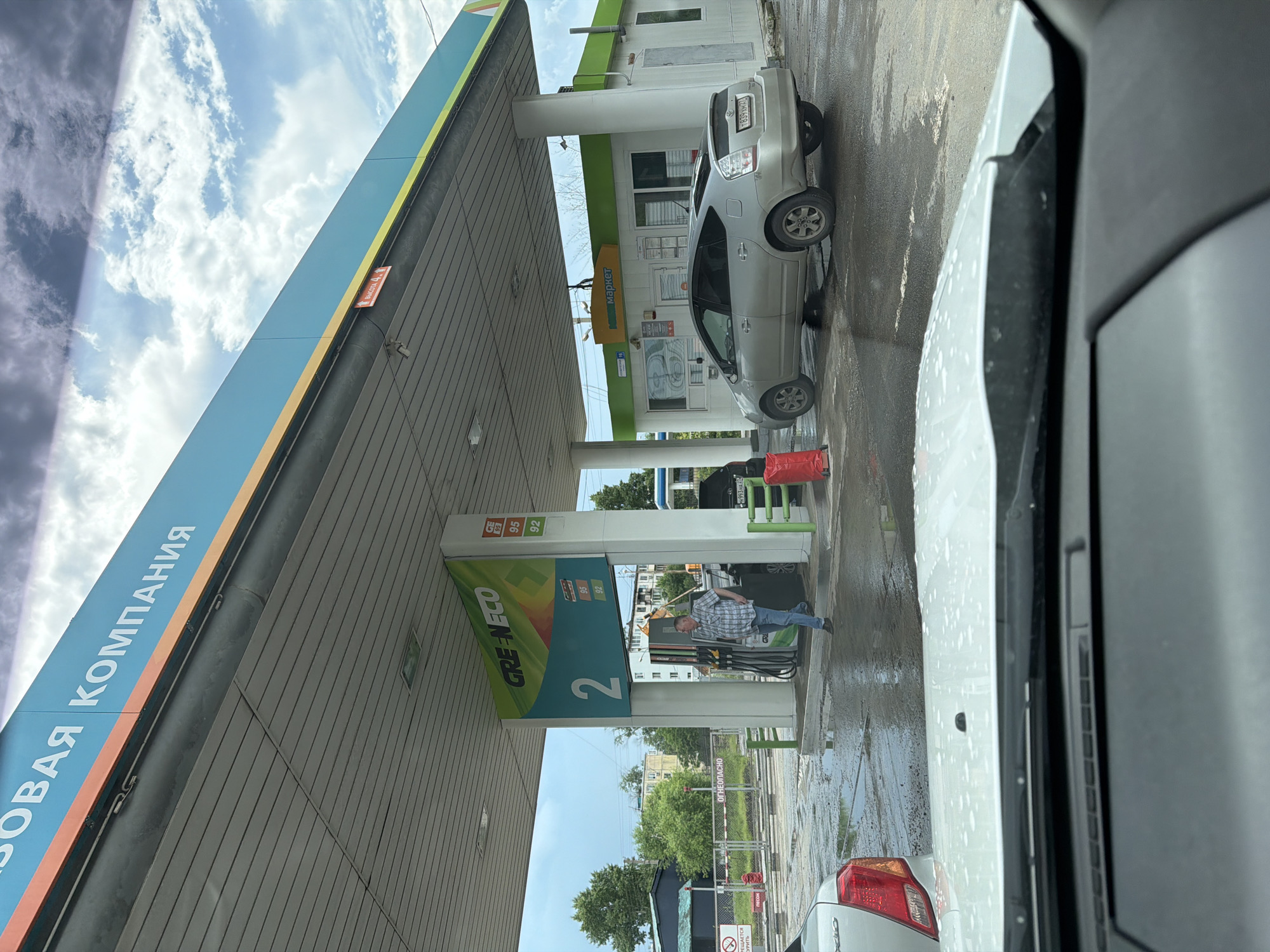 The image shows a gas station in Khabarovsk, Russia. A man in a plaid shirt is standing at a gas pump. A silver Toyota Prius is parked at another pump. The ground is wet, suggesting it has recently rained. There are several buildings in the background, including a convenience store labeled "Supermarket." The sky is cloudy.