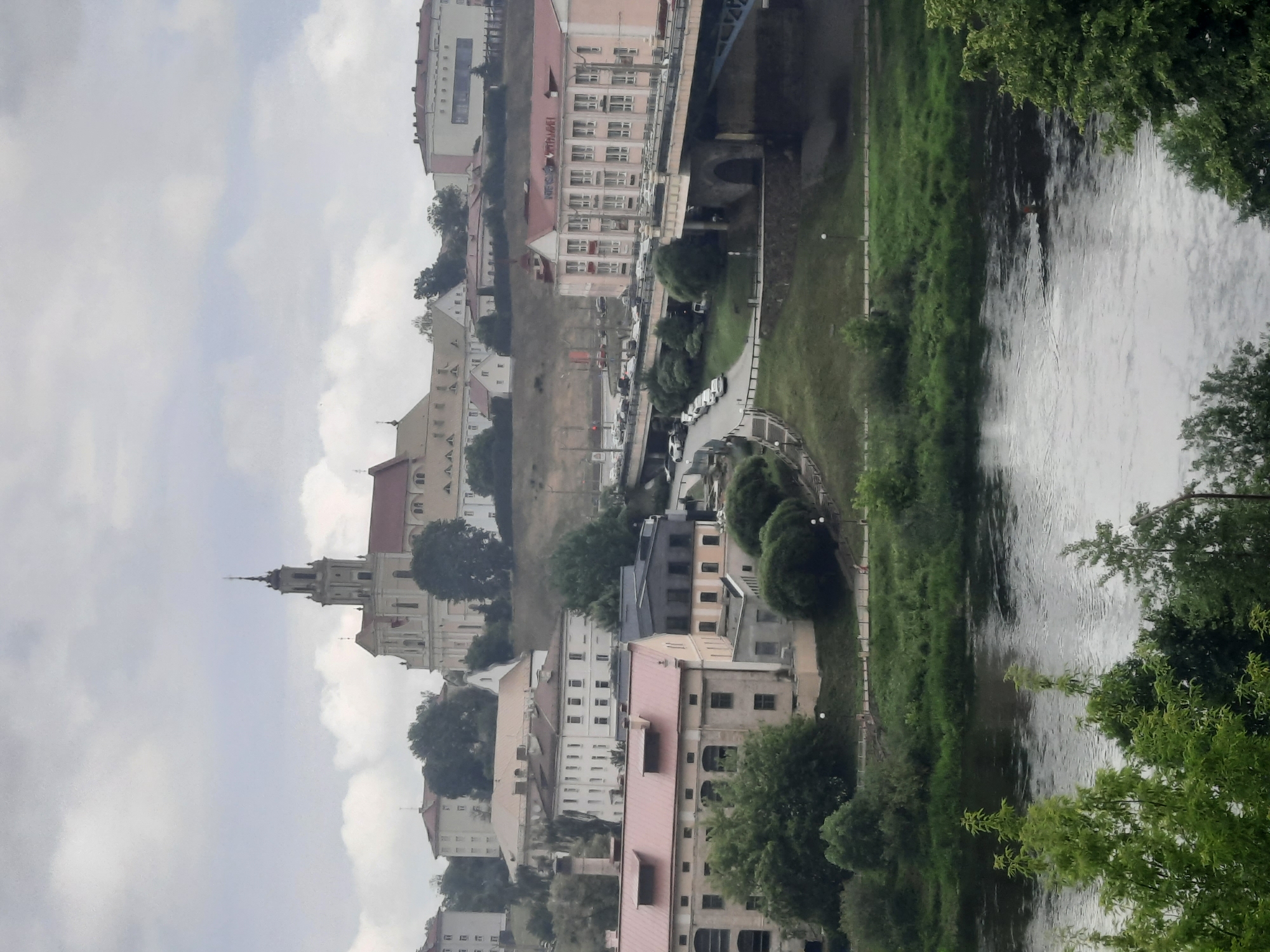 The image shows a panoramic view of Hrodna, Belarus, featuring a river in the foreground and a cityscape in the background. The river, likely the Neman River, flows gently, with a person swimming or wading in its water. The riverbank is lined with lush green grass and trees.
Dominating the background is a large, light-colored church with a tall spire, believed to be the Farny Church. Adjacent to it are other historical buildings and residential structures. A multi-lane bridge, likely the Old Bridge or a section of the Hrodna ring road, spans across the river, with numerous cars visible on it and on the roads leading to it. The overall atmosphere is overcast, contributing to a muted color palette. The image captures a blend of natural scenery and urban development, showcasing the distinct architectural style of Hrodna.
