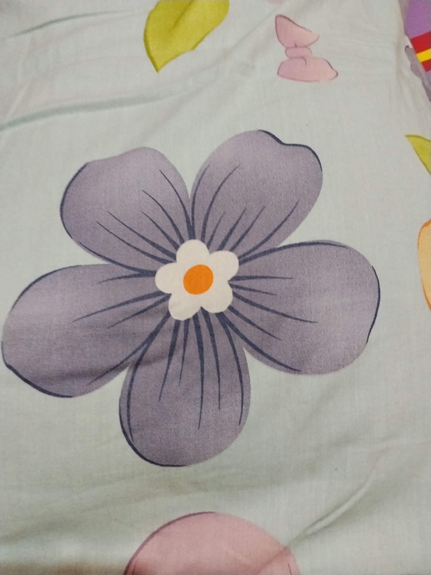 The image shows a close-up of a fabric with a floral pattern. A large, gray flower with five petals is centered in the frame. The flower has a small, white center with an orange dot in the middle. Dark gray lines radiate from the center of the flower along each petal. The background fabric is a light pastel green color, and there are other, smaller floral and abstract shapes scattered around the main flower, including a yellow leaf, a pink shape resembling a bow, and a portion of a pink circular shape. The location is indeterminable, and no people, subjects, objects, actions, or interactions can be definitively identified.