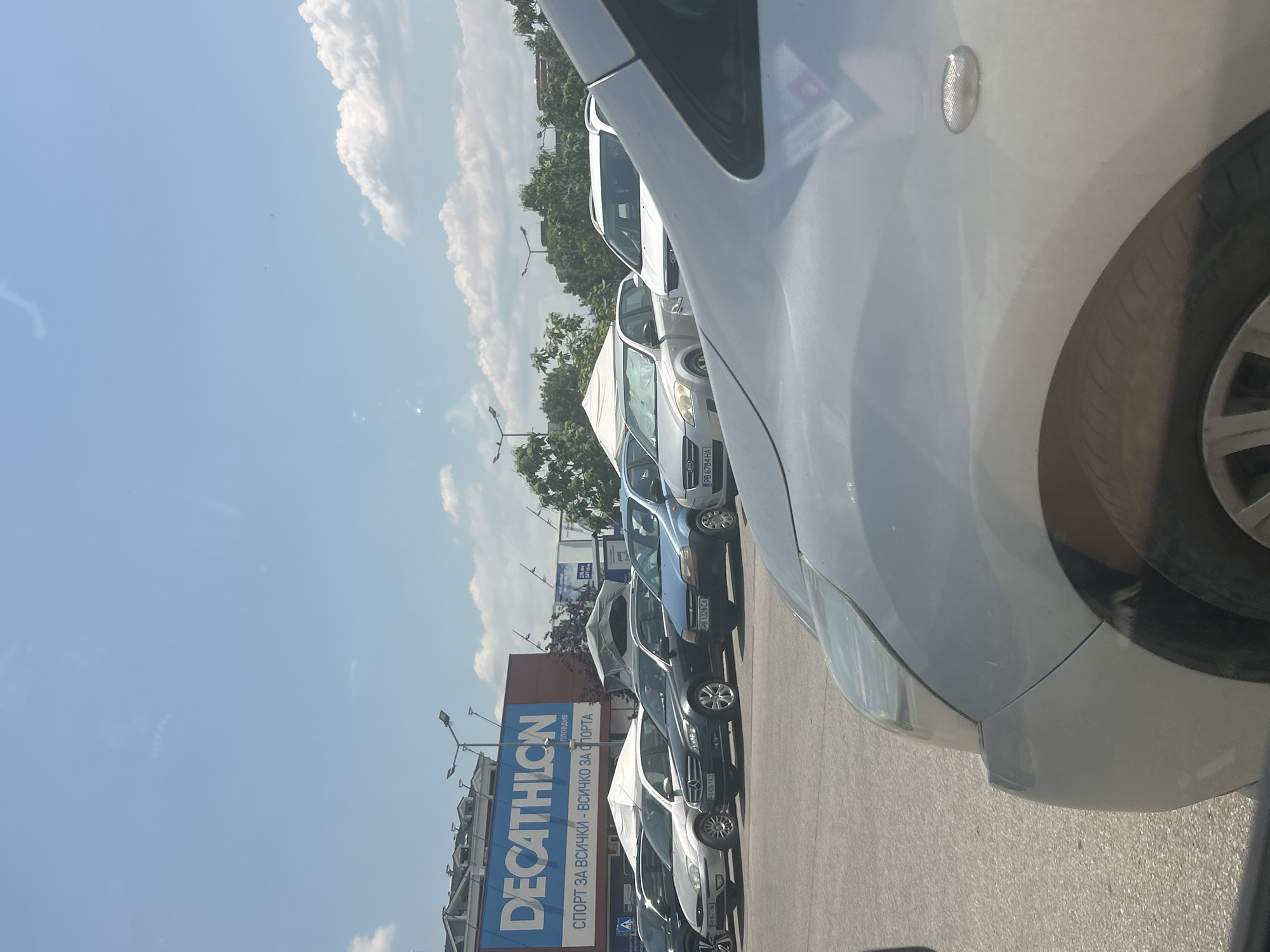 The image depicts a parking lot outside a Decathlon store in Plovdiv, Bulgaria. The store's large blue sign with white lettering is prominently displayed. Several cars are parked in the lot, with a silver car in the foreground partially obscuring the view. The sky is blue with scattered clouds, and trees and streetlights are visible in the background. The scene appears to be captured during daylight hours.