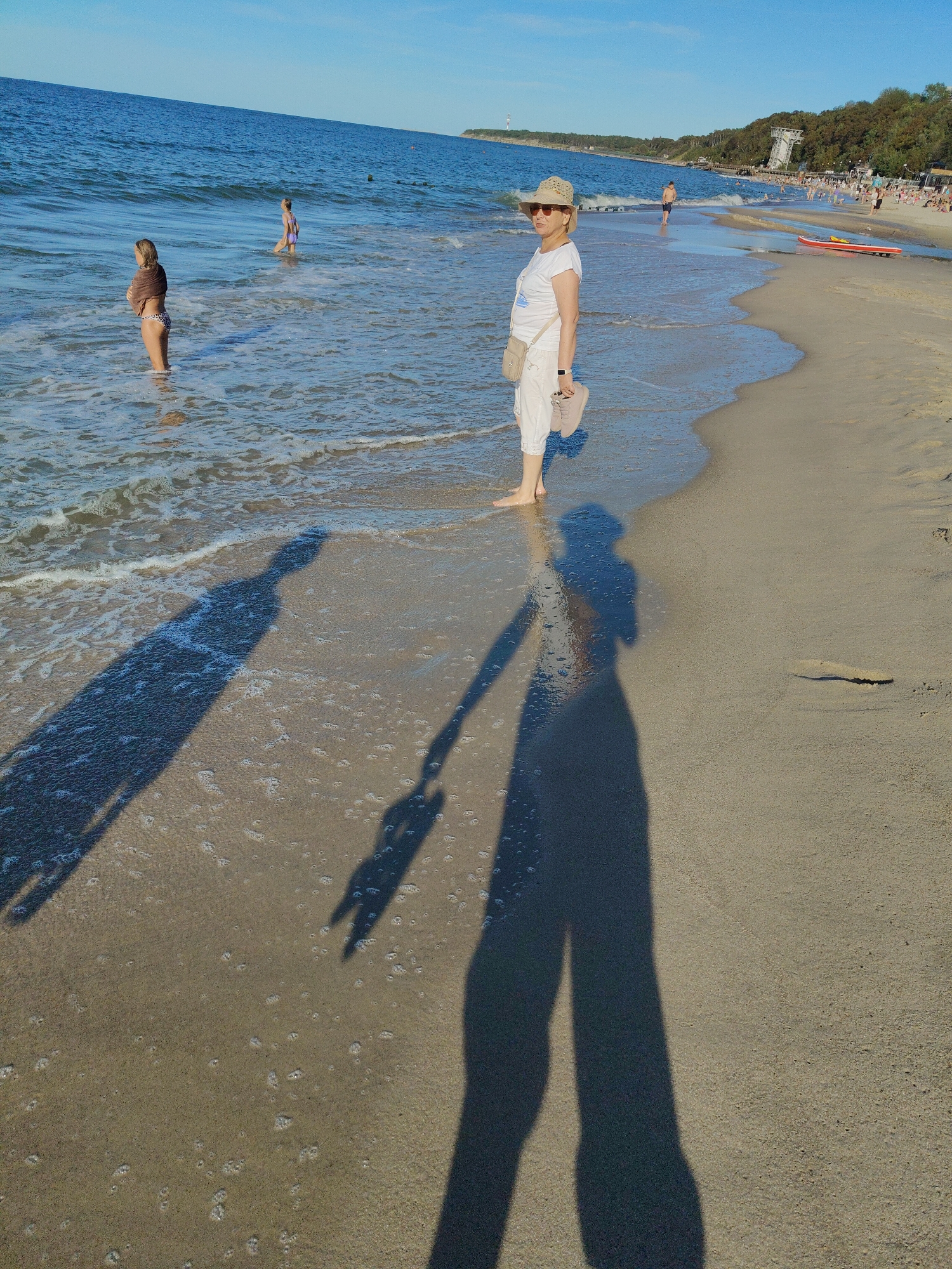 The image depicts a sunny day at a beach, likely in Pionersky, Russia, given the presence of the Pionersky Lighthouse in the background. In the foreground, a woman wearing a white t-shirt, white capri pants, and a straw hat stands at the edge of the water, holding a pair of beige shoes. Her shadow stretches across the wet sand. Two children are visible in the water to the left; one, wearing a leopard-print bikini bottom and a brown wrap, is facing away from the camera, while another child in a purple bikini is further out in the sea. In the mid-ground, other beachgoers are visible, some in the water and some on the sand. A red kayak lies on the beach near the water's edge, and a light-colored building with a tall structure stands on a hill in the distance, identified as the Pionersky Lighthouse. The sea is calm with small waves lapping at the shore, and the sky is clear and blue. The sand is a light brown, and the overall atmosphere is one of leisure and recreation.