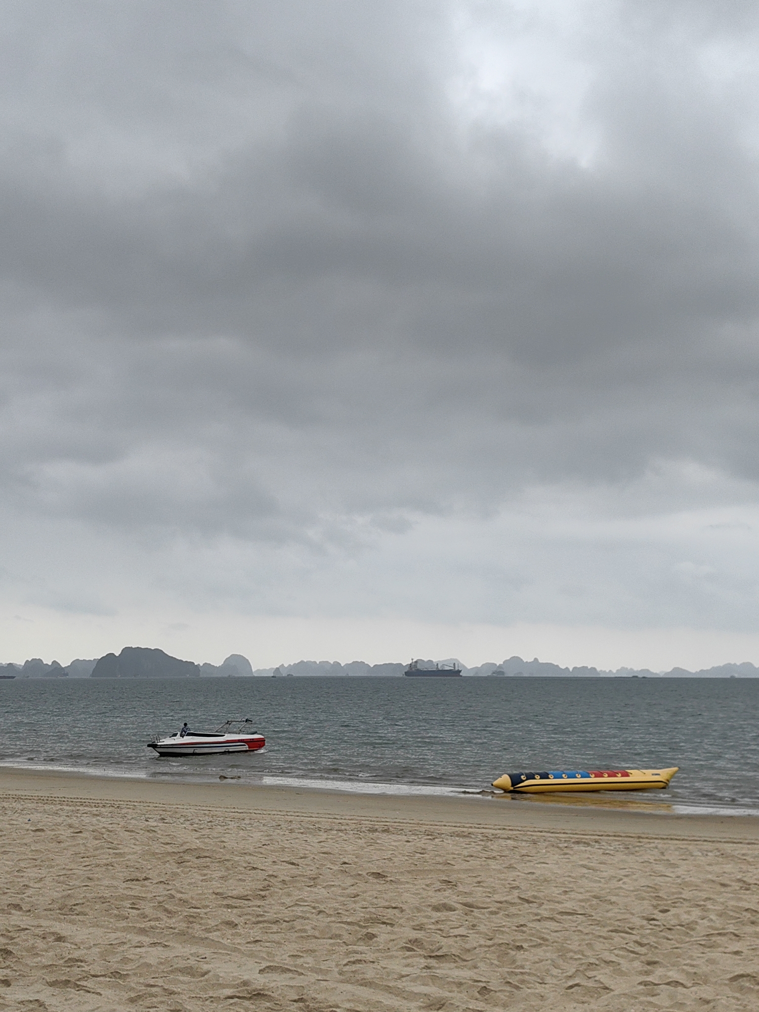 The image depicts a beach scene in Bai Chay Ward, Vietnam, under a heavy, overcast sky. The foreground features a sandy beach. The water line separates the beach from the gray sea, where a motorboat and a banana boat rest on the shore. The banana boat is predominantly yellow with blue and red accents. The motorboat is white with red detailing. In the distance, there are blurred outlines of rocky islands or land formations, with a large cargo ship visible on the left side.