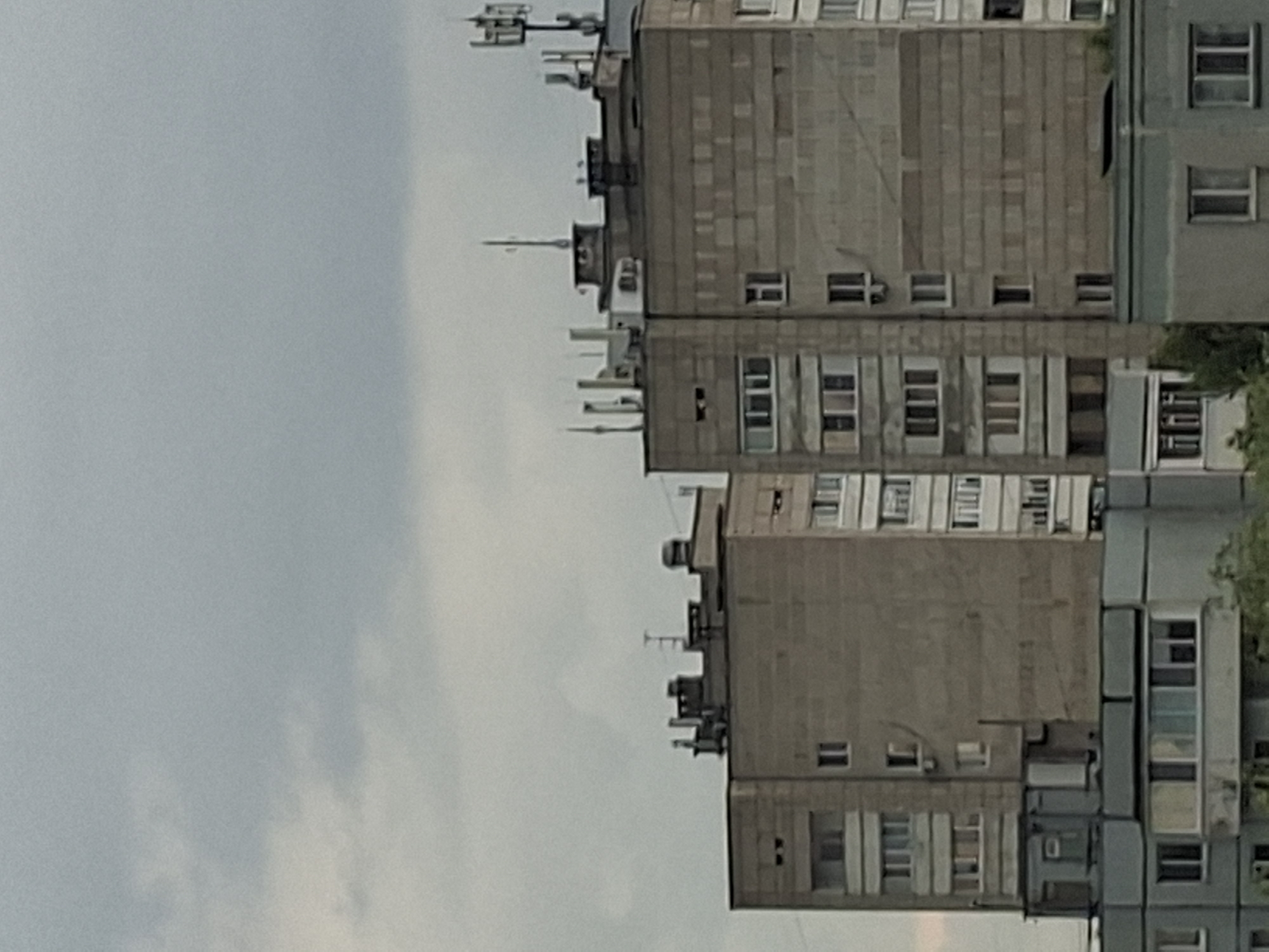 The image shows a cloudy sky over a view of apartment buildings in Kazan, Russia. The buildings are predominantly concrete, with visible brickwork on some facades. There are multiple antennas and communication equipment mounted on the roofs of the taller buildings, suggesting an urban environment. The lower parts of the buildings show balconies and numerous windows. The scene appears to be a typical residential area, captured from a distance, possibly from another building or a slightly elevated viewpoint. No people are visible in the image.