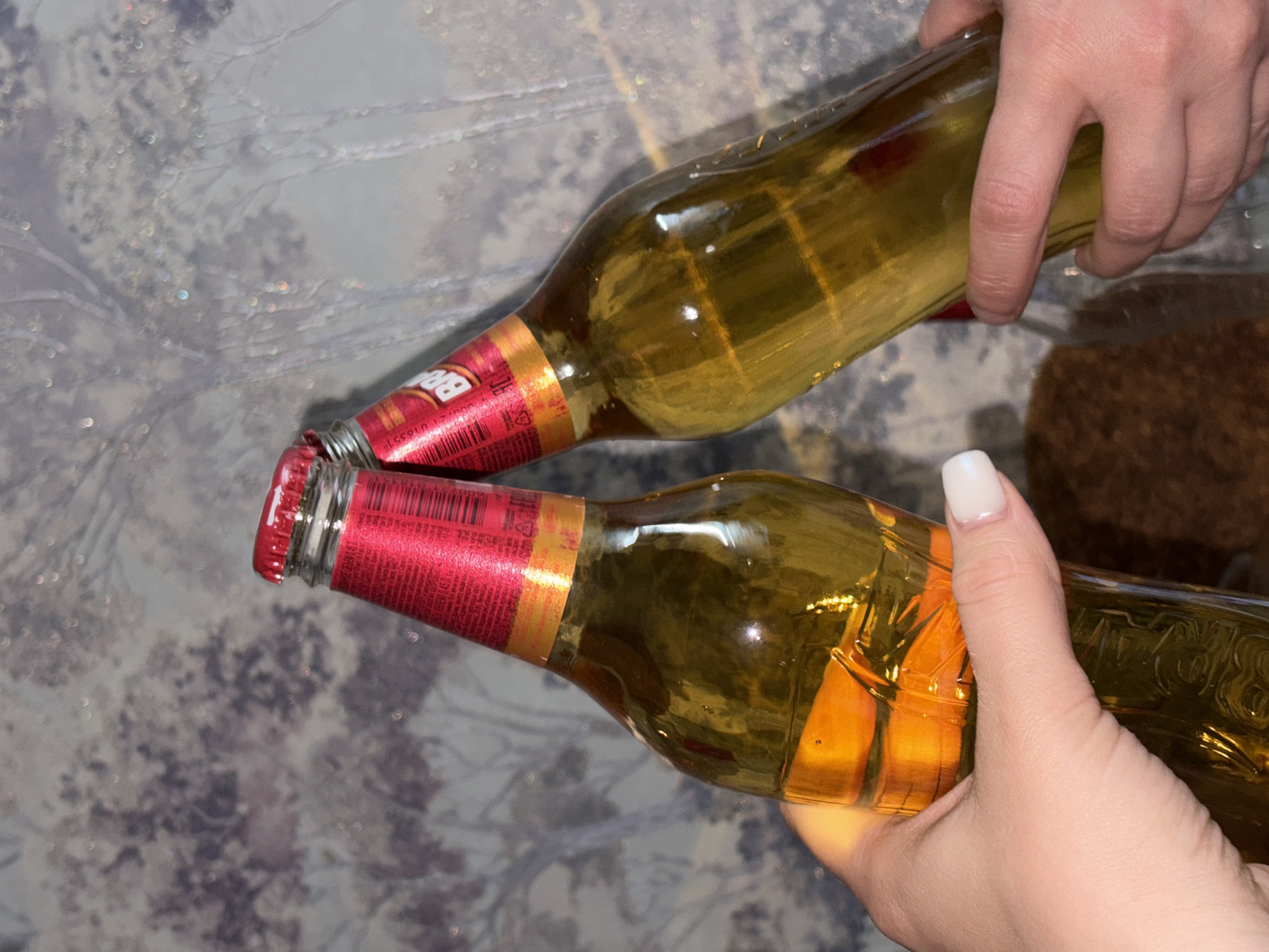 Two hands hold two brown glass beer bottles, appearing to clink the bottles together at the necks. The bottles have red caps and labels. The background is a blurred surface with a mottled pattern. There is no verifiable information in the image to confirm its location as Novopokrovskaya, Russia.