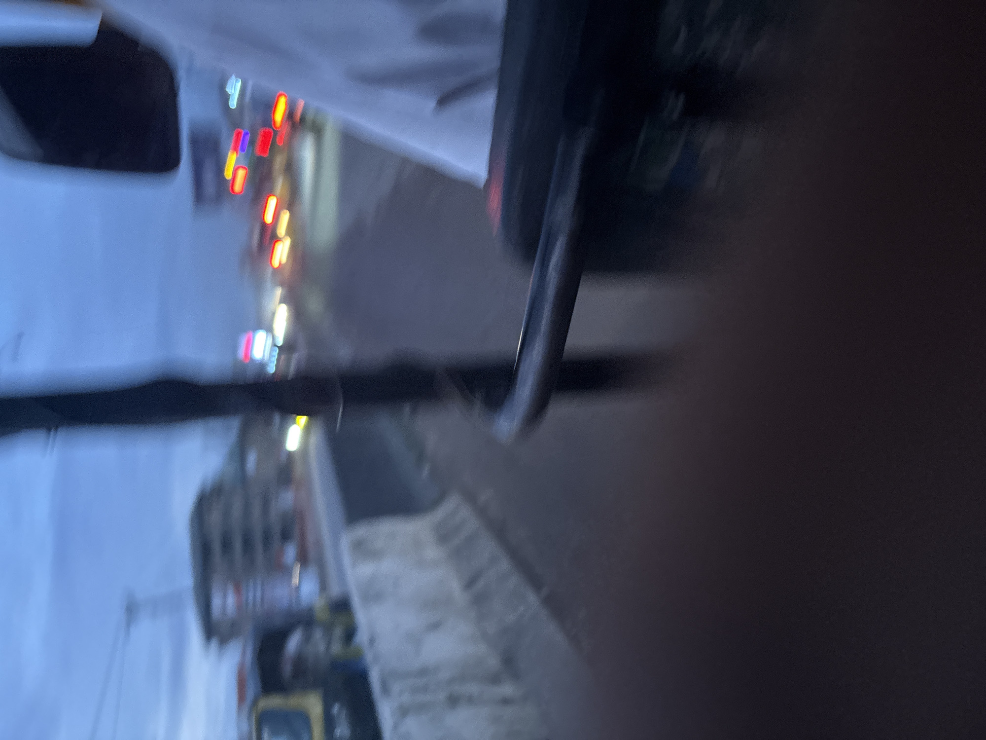 The image depicts a blurred street scene at dusk or dawn in Onitsha, Nigeria. In the foreground, a dark, blurry object, possibly a railing or part of a vehicle, is visible. To the left, a portion of a yellow vehicle is seen. A concrete barrier runs along the side of the road. In the background, buildings and trees are indistinct. Streaks of light from moving vehicles are evident, suggesting traffic on the road. The sky is overcast with a blue-grey hue. The overall impression is one of motion and low visibility, characteristic of a busy urban environment during low-light conditions.