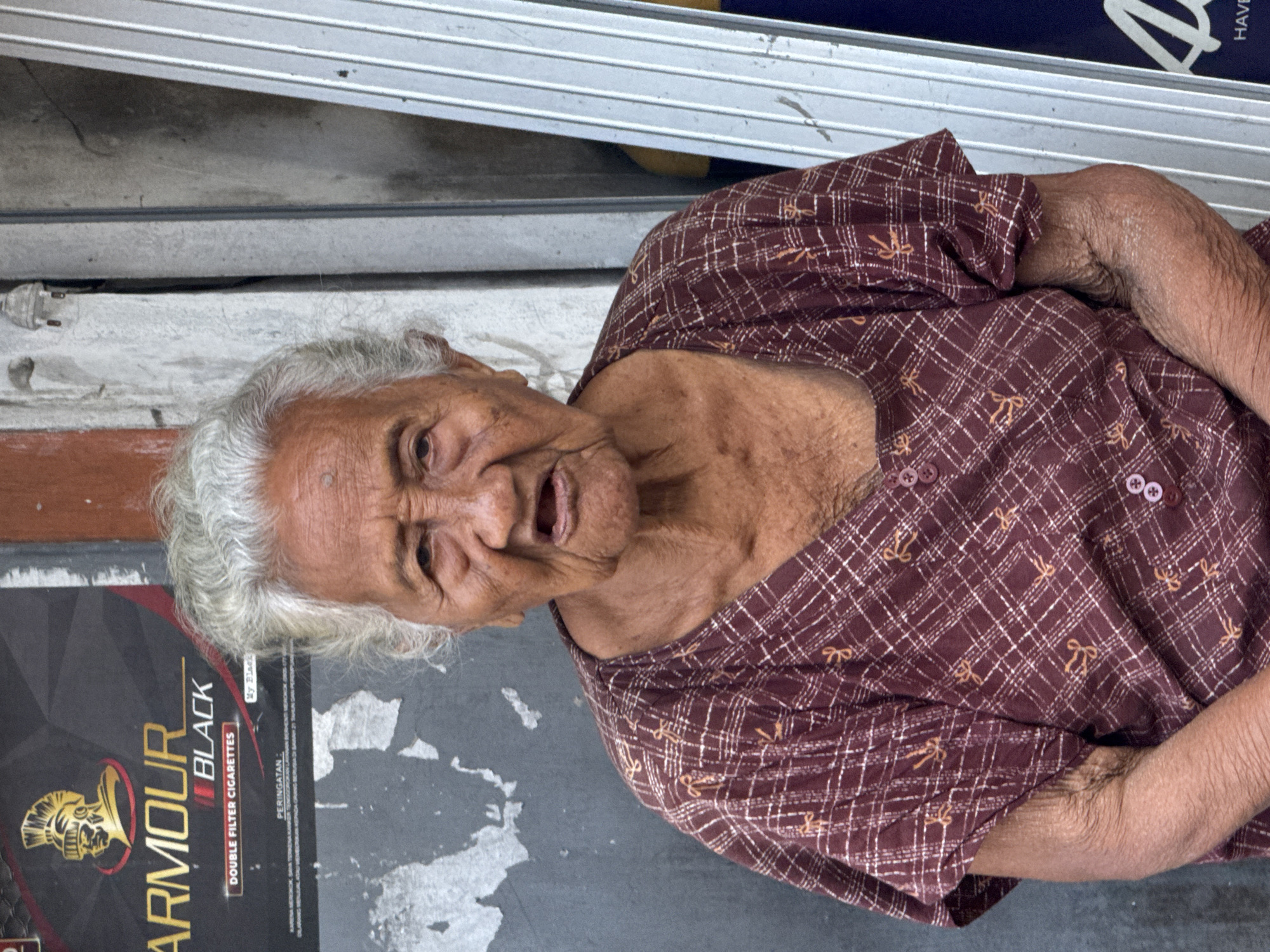 An elderly woman with grey hair is seated on the ground next to a cigarette advertisement with the text "ARMOUR BLACK DOUBLE FILTER CIGARETTES". She is wearing a maroon plaid shirt. The background appears to be a building exterior with a corrugated metal door. The location is Kuta, Indonesia.