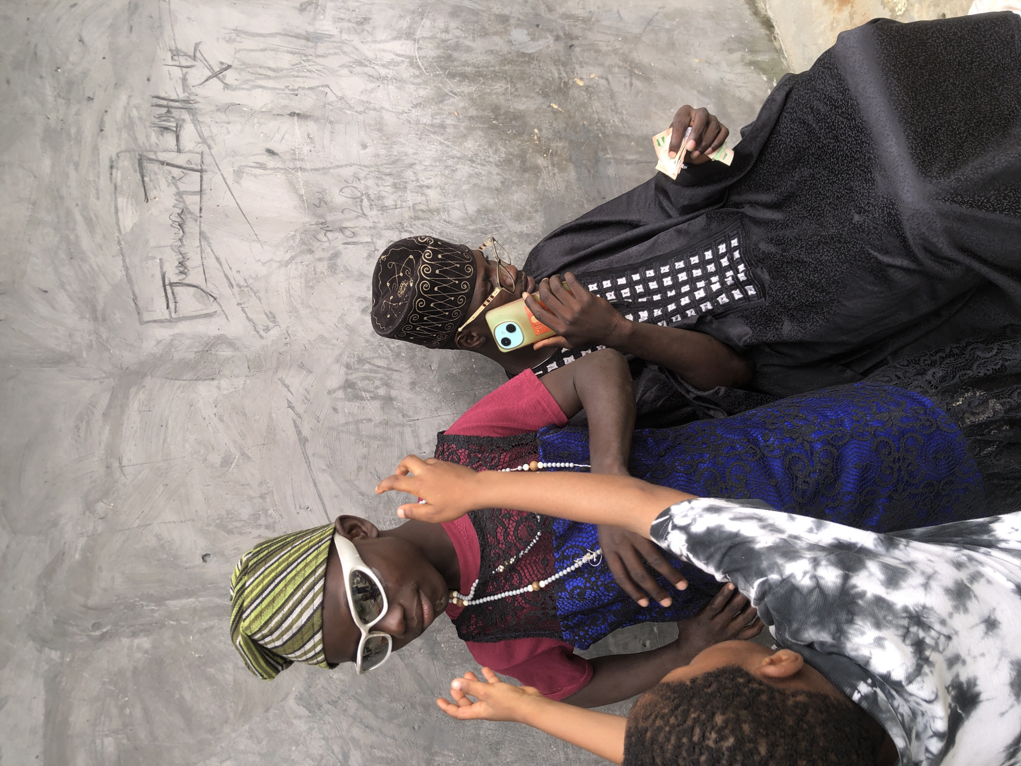 Three young Black males are captured outdoors in Ado, Nigeria, against a textured grey concrete wall with faint graffiti. The central figure, wearing a green and white striped cap and white-rimmed sunglasses, is adorned with a burgundy t-shirt layered under a blue lace dress and multiple beaded necklaces. His gaze is directed downward. To his right, another young male, dressed in a black embroidered cap and traditional black attire, is seated and holding a light-colored smartphone in his right hand while his left hand clutches a small fan of currency. A third boy, partially visible in the foreground, faces away from the camera, his back and arms clad in a black and white tie-dye shirt. His hands are raised as if interacting with the central figure. The environment appears to be a sparsely furnished outdoor space, possibly a courtyard or alleyway, with the concrete wall forming the backdrop.