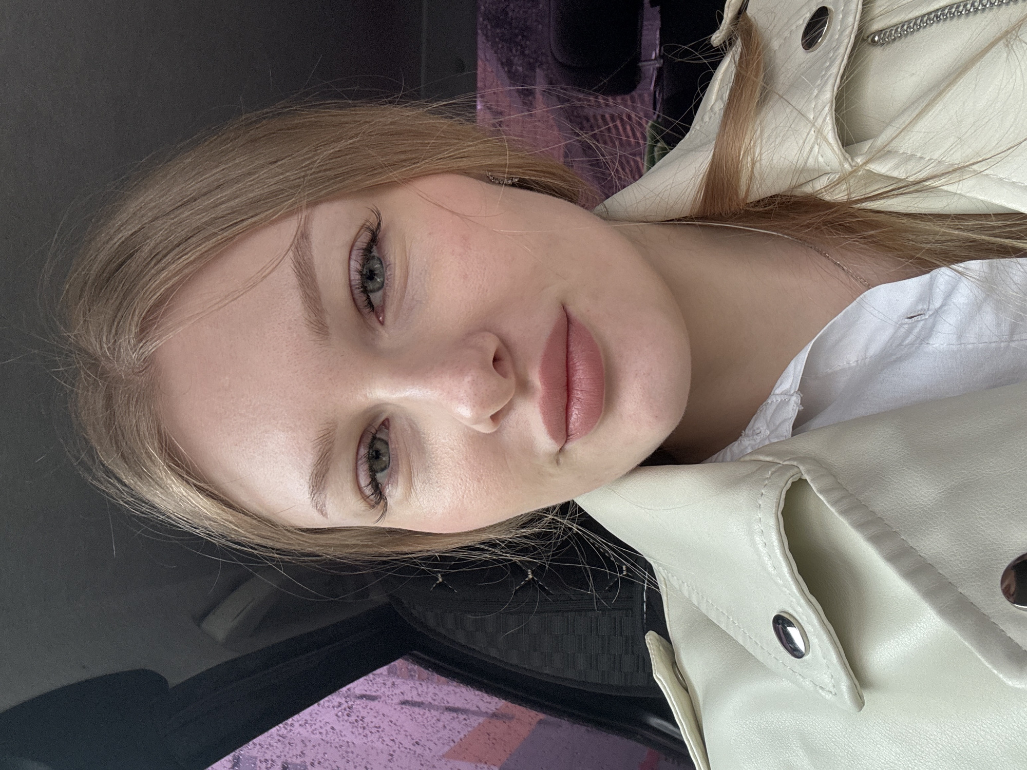 A young Caucasian woman with blonde hair and light green eyes is depicted in a close-up shot. She is wearing a white jacket over a white shirt, a silver chain on her neck. The background shows a car interior, the street visible through the car window has a pinkish and purple tones. The woman is looking at the camera. The scene appears to be indoors, in the car. There is no data available on the precise location of the city of Perm, Russia, in the image.