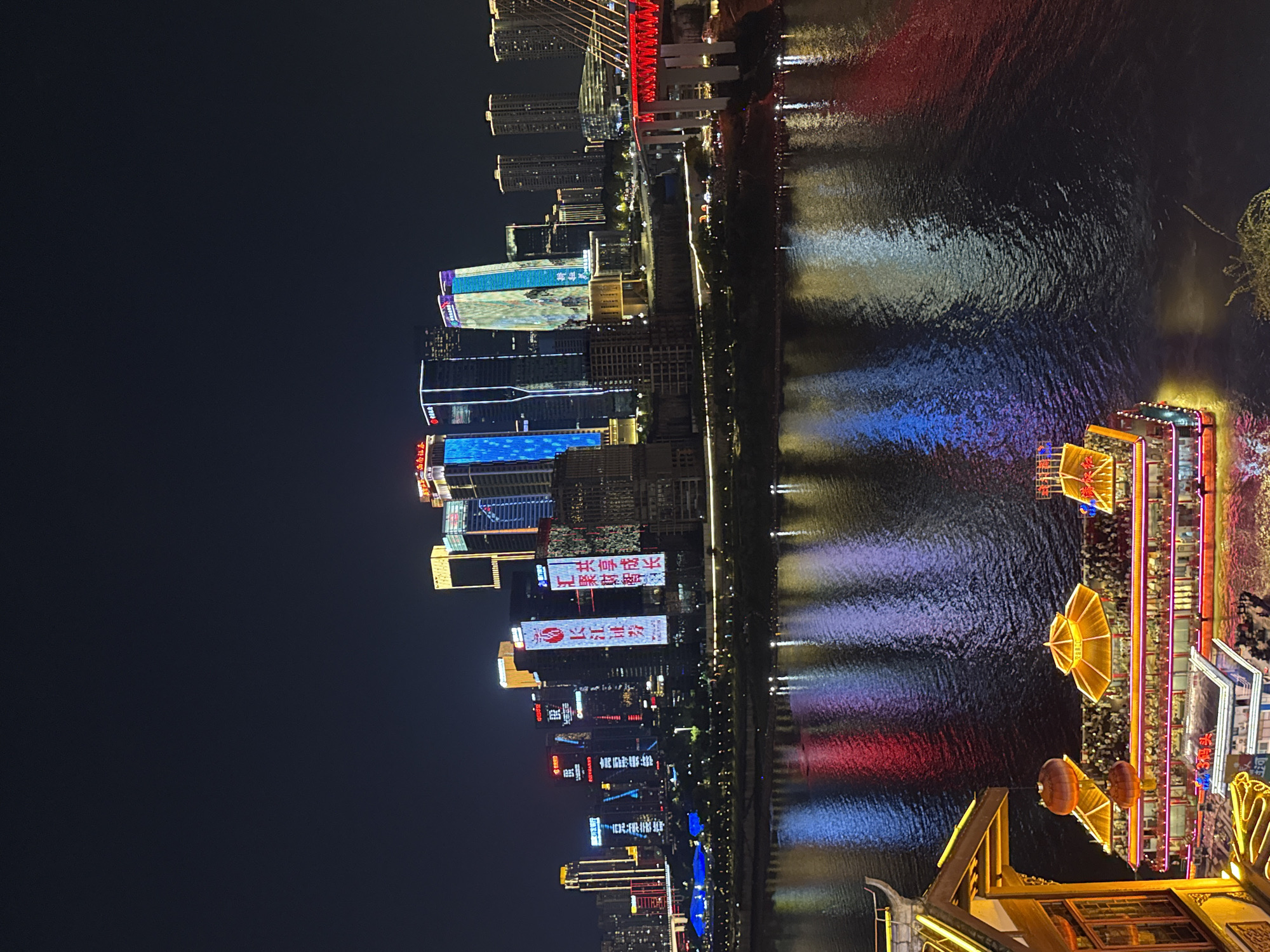 This is a nighttime aerial shot of the Yuzhong District in Chongqing, China, focusing on the Jialing River and the illuminated cityscape. On the left, a traditional Chinese-style building with red lanterns and a golden roof is partially visible in the foreground, jutting out over the water. A large, multi-decked boat, adorned with yellow canopies and colorful lights, is docked and crowded with people. The river reflects the vibrant lights of the city, creating streaks of red, blue, and white on its dark surface. In the background, a dense collection of modern skyscrapers dominates the skyline, many featuring illuminated signs and building facades, showcasing a bustling urban environment. A large, red-lit bridge spans the river in the right portion of the image, further emphasizing the city's infrastructure and nighttime activity. The sky is dark, indicating the late hour.